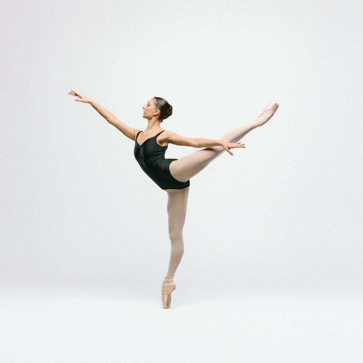 Dancer in black leotard executes arabesque on pointe against white background.