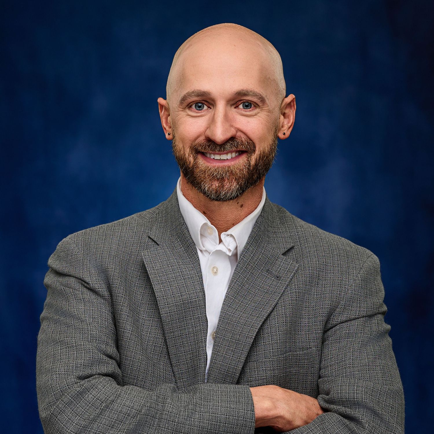 Professional headshot of bald man with beard in grey suit against blue backdrop.