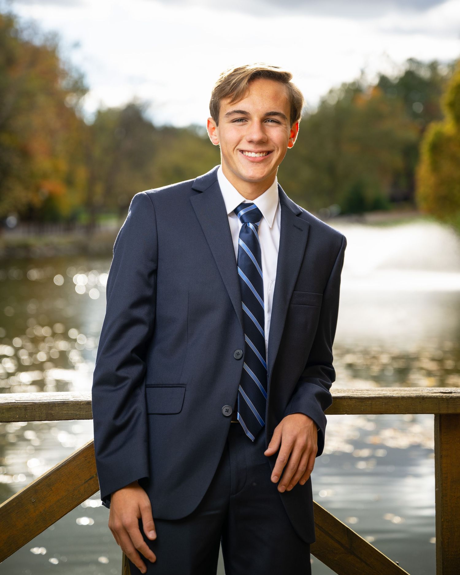 Professional portrait of young man in navy suit on deck by lake.