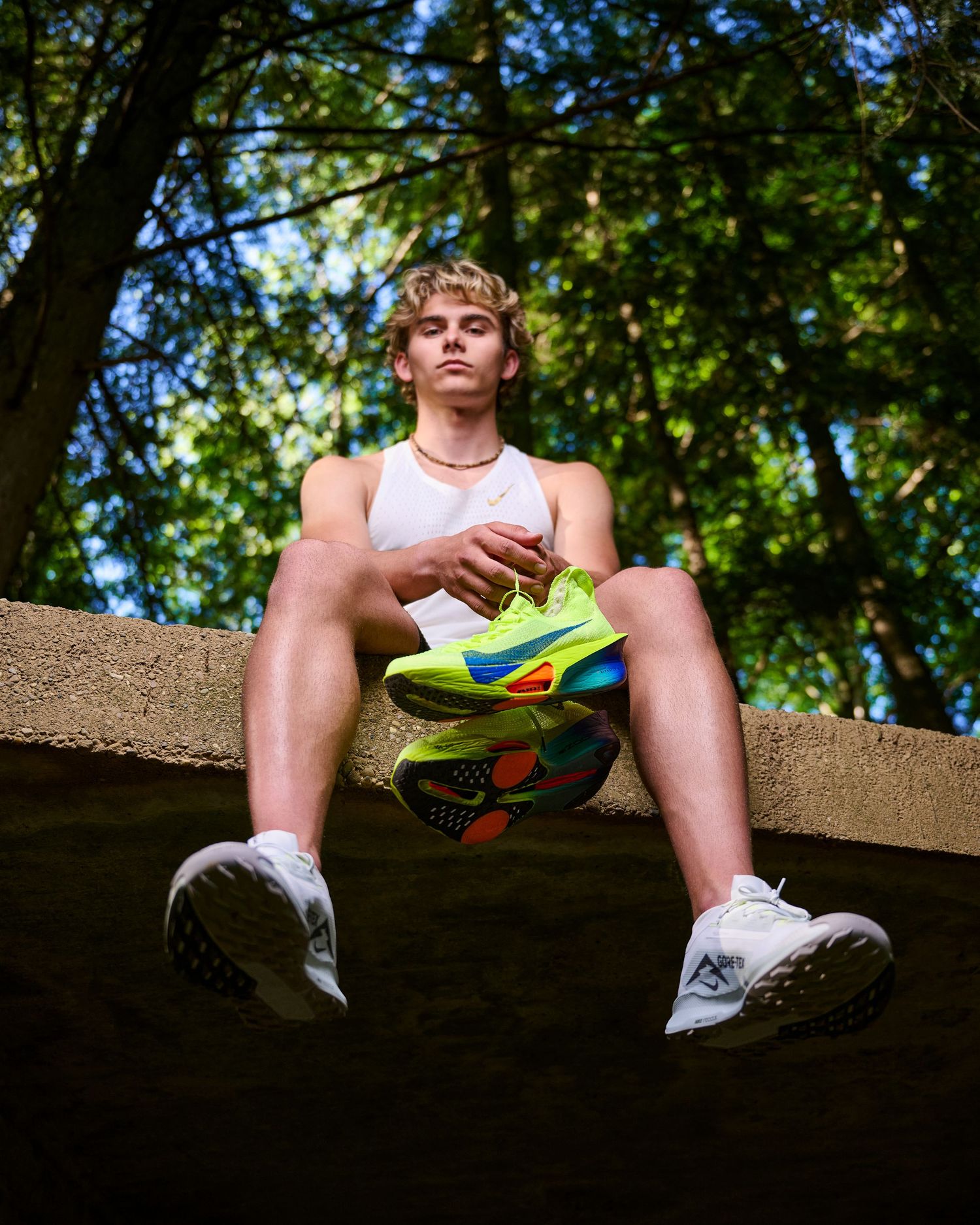 Person sitting on a concrete ledge holding colorful athletic shoes in a lush green outdoor setting.