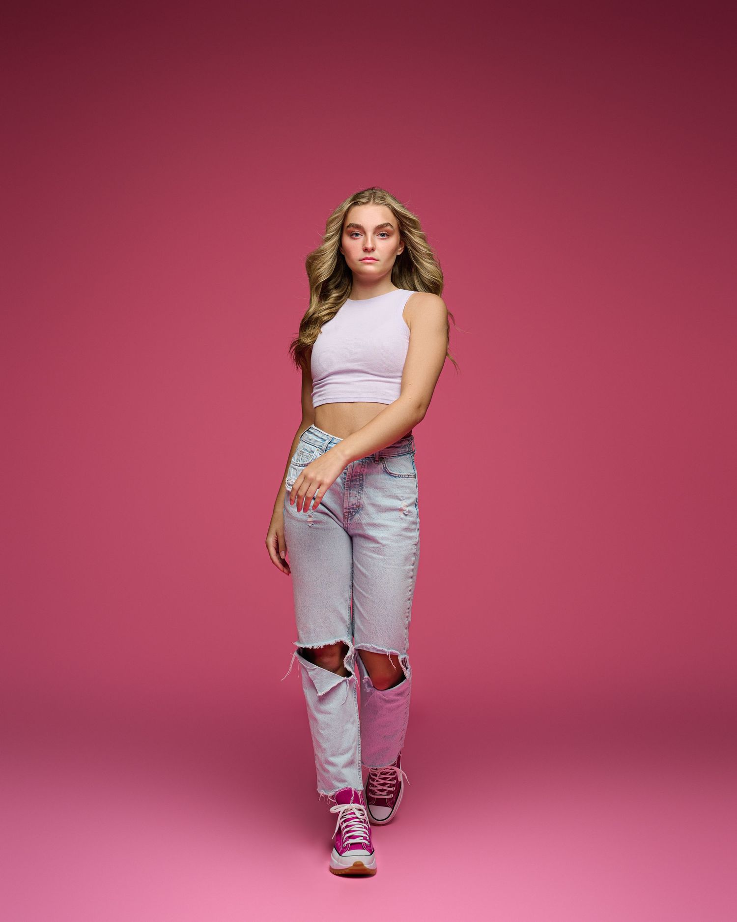 Model in crop top and ripped jeans poses against pink backdrop.