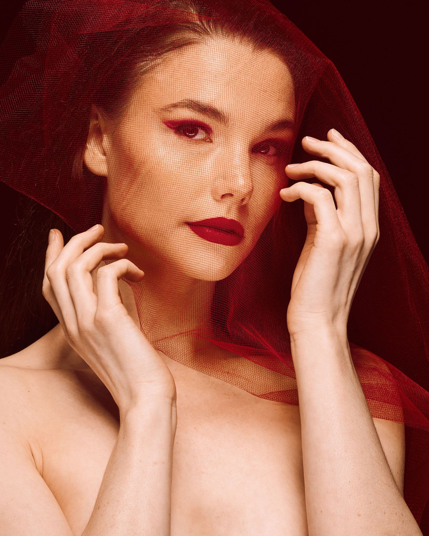 Beauty portrait with sheer red veil and dramatic makeup on crimson background.