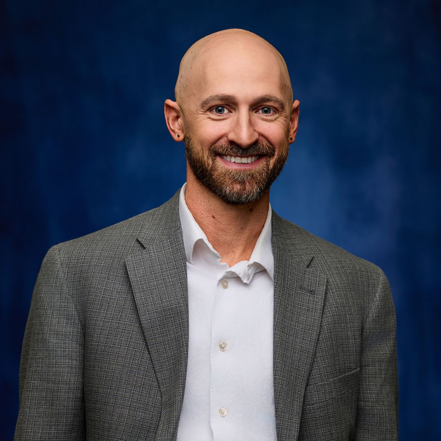 Professional headshot of person in gray suit jacket and white shirt against blue background.