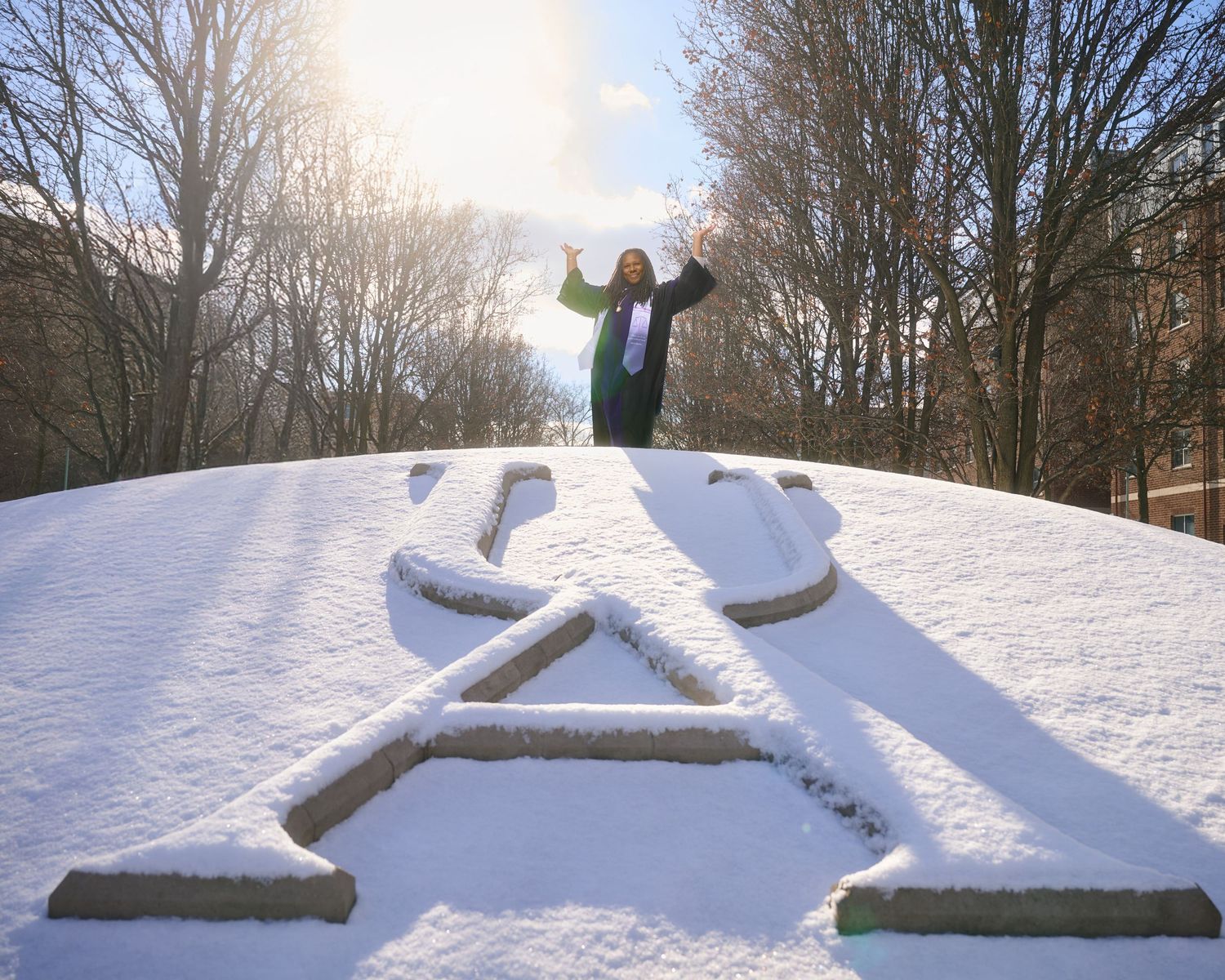 Graduate celebrates on snow-covered university emblem during winter.