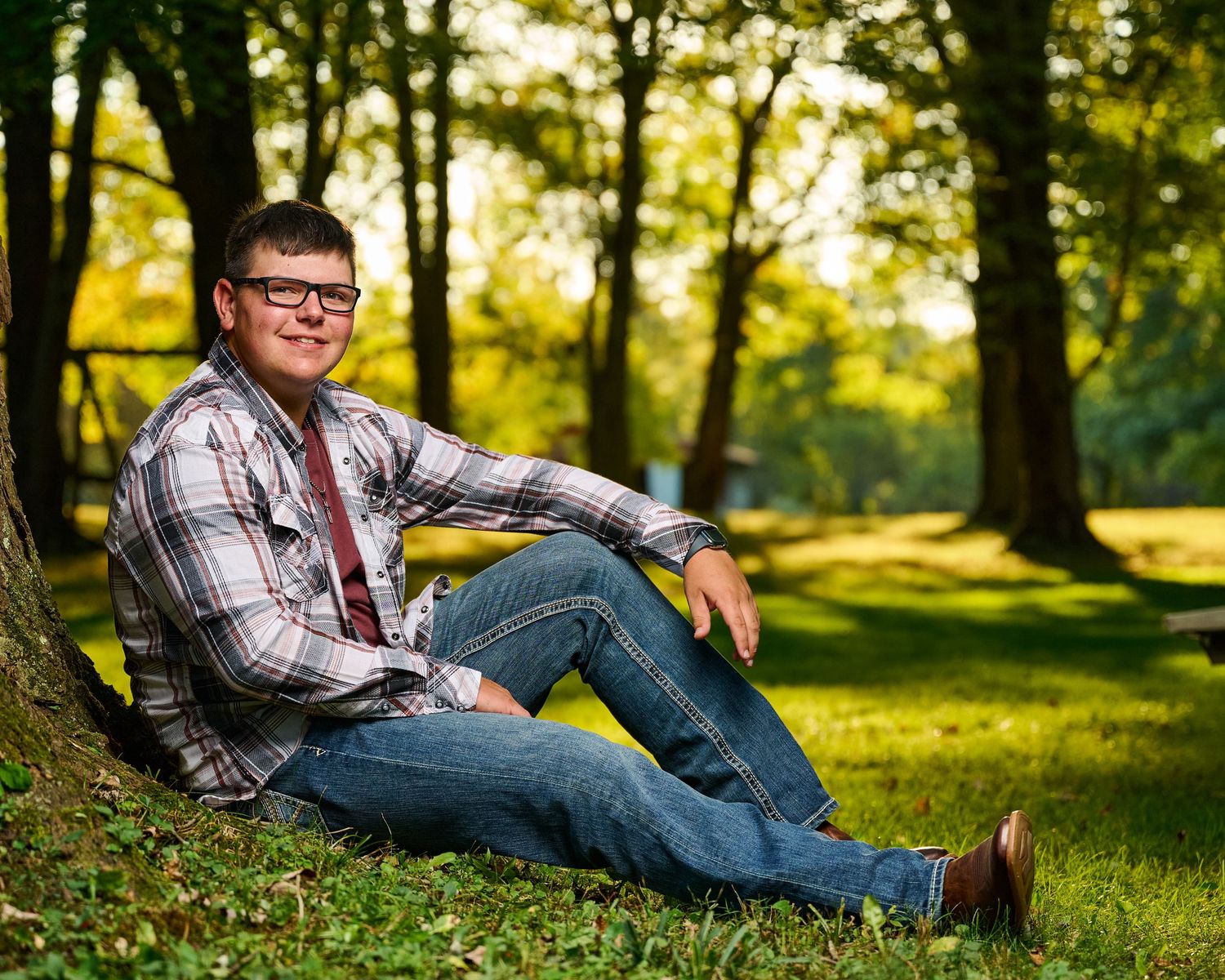A casual outdoor portrait of someone in plaid and jeans relaxing against a tree in a sunlit park.