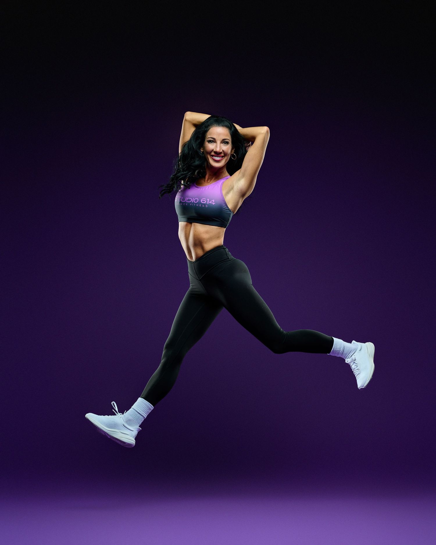 Athletic woman in black activewear and white sneakers jumps energetically against a dark purple background.