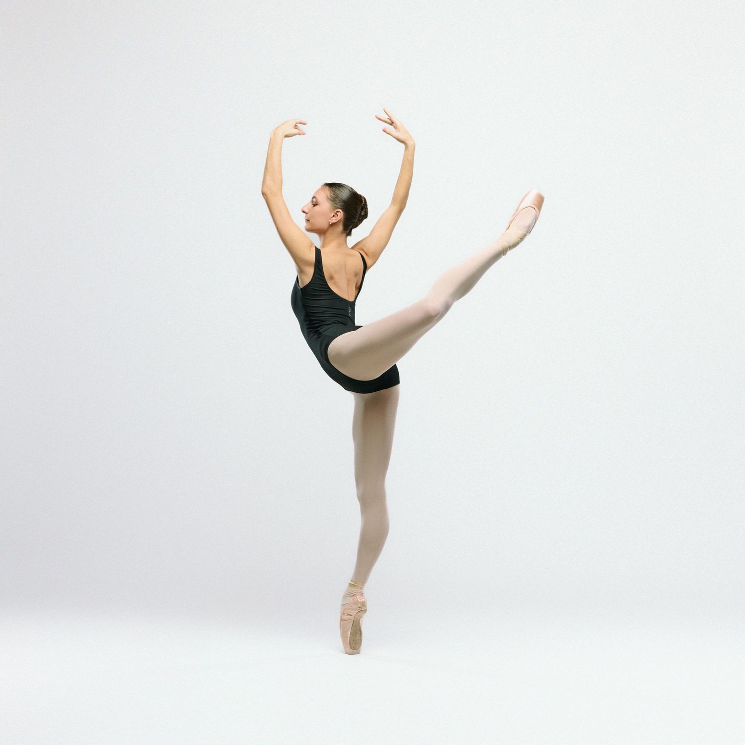 Ballet dancer performs graceful arabesque en pointe in black leotard against white backdrop.