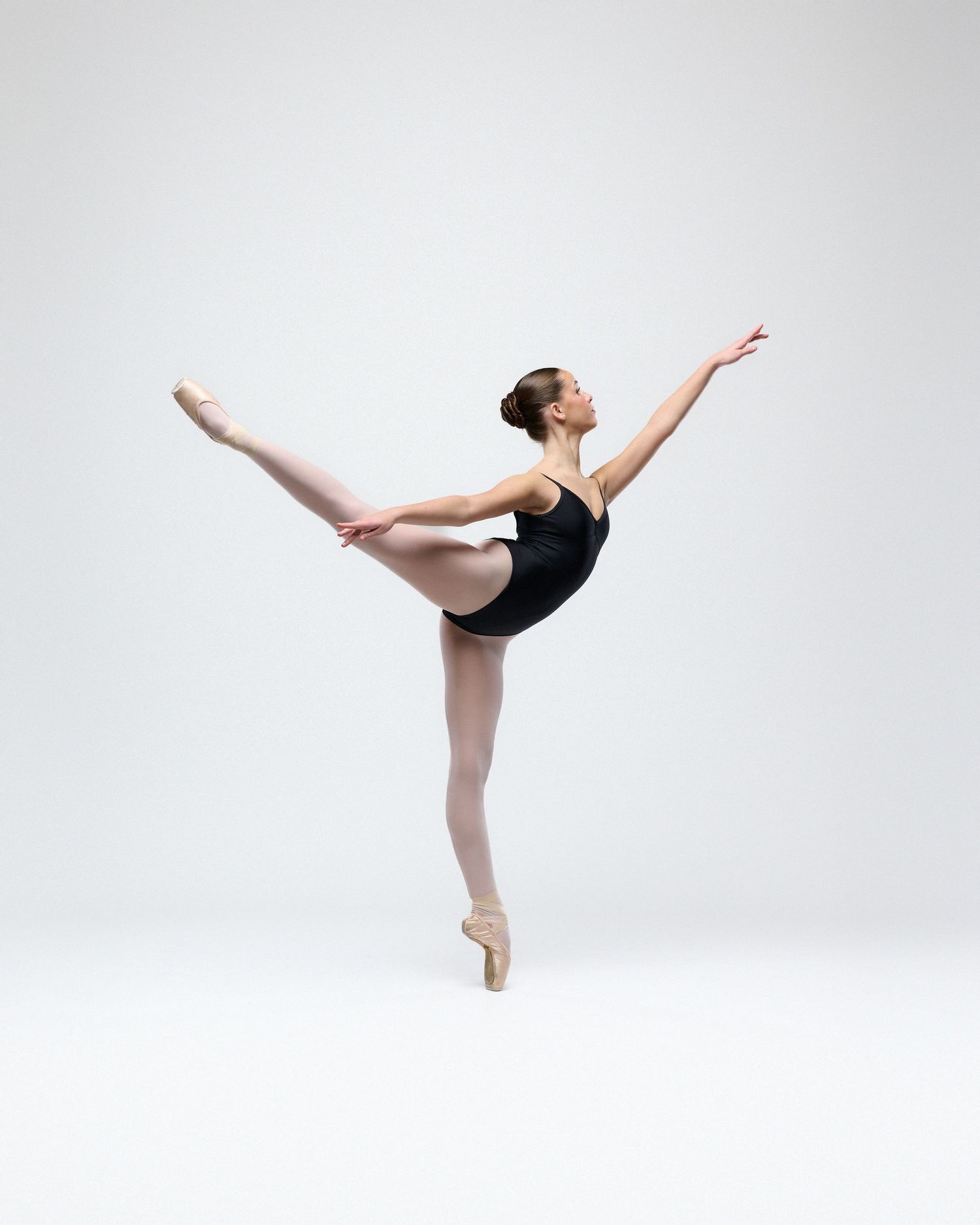 Ballet dancer in black leotard executes graceful arabesque position against white backdrop.