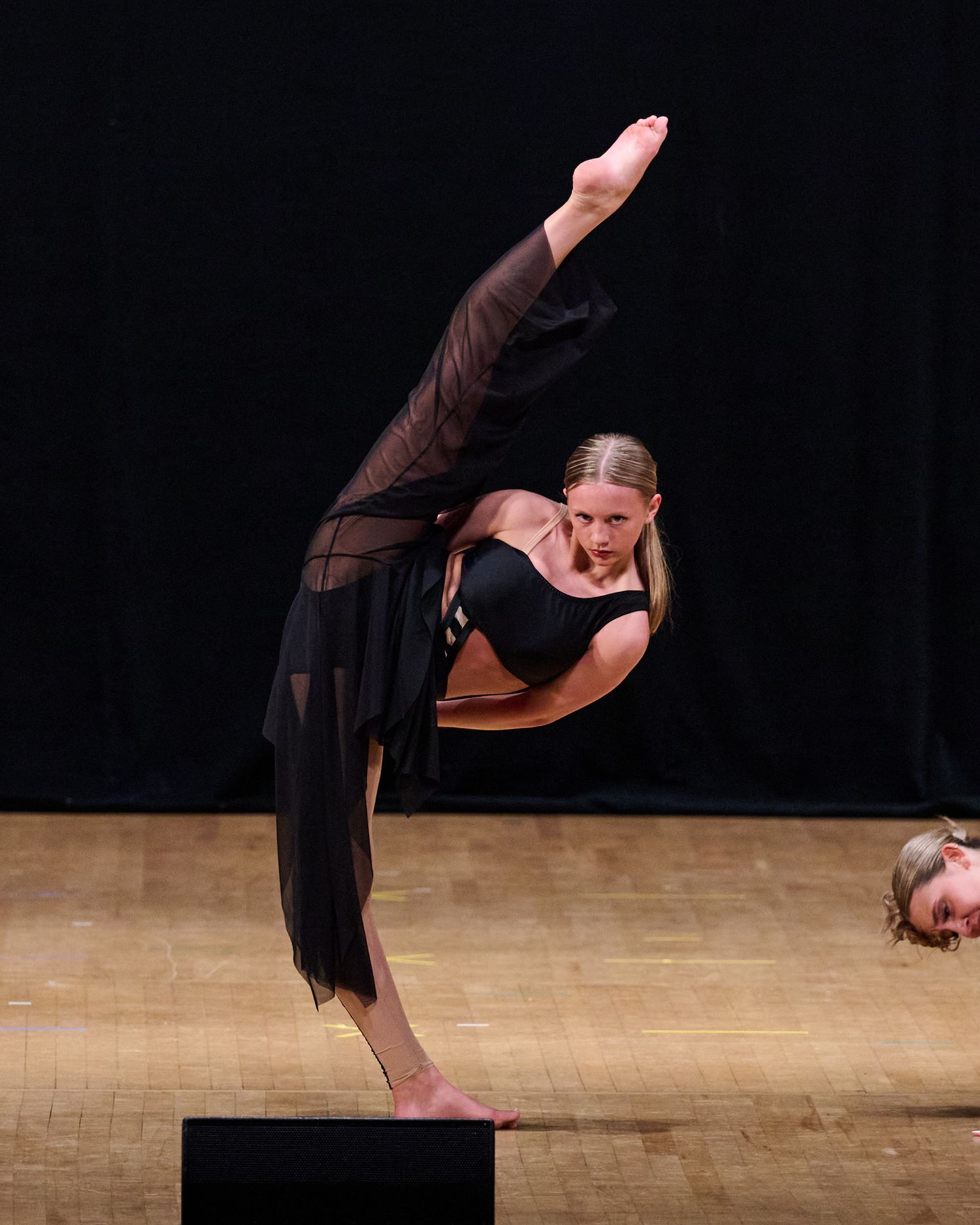 A dancer in black performs a dramatic high kick on stage during a performance.