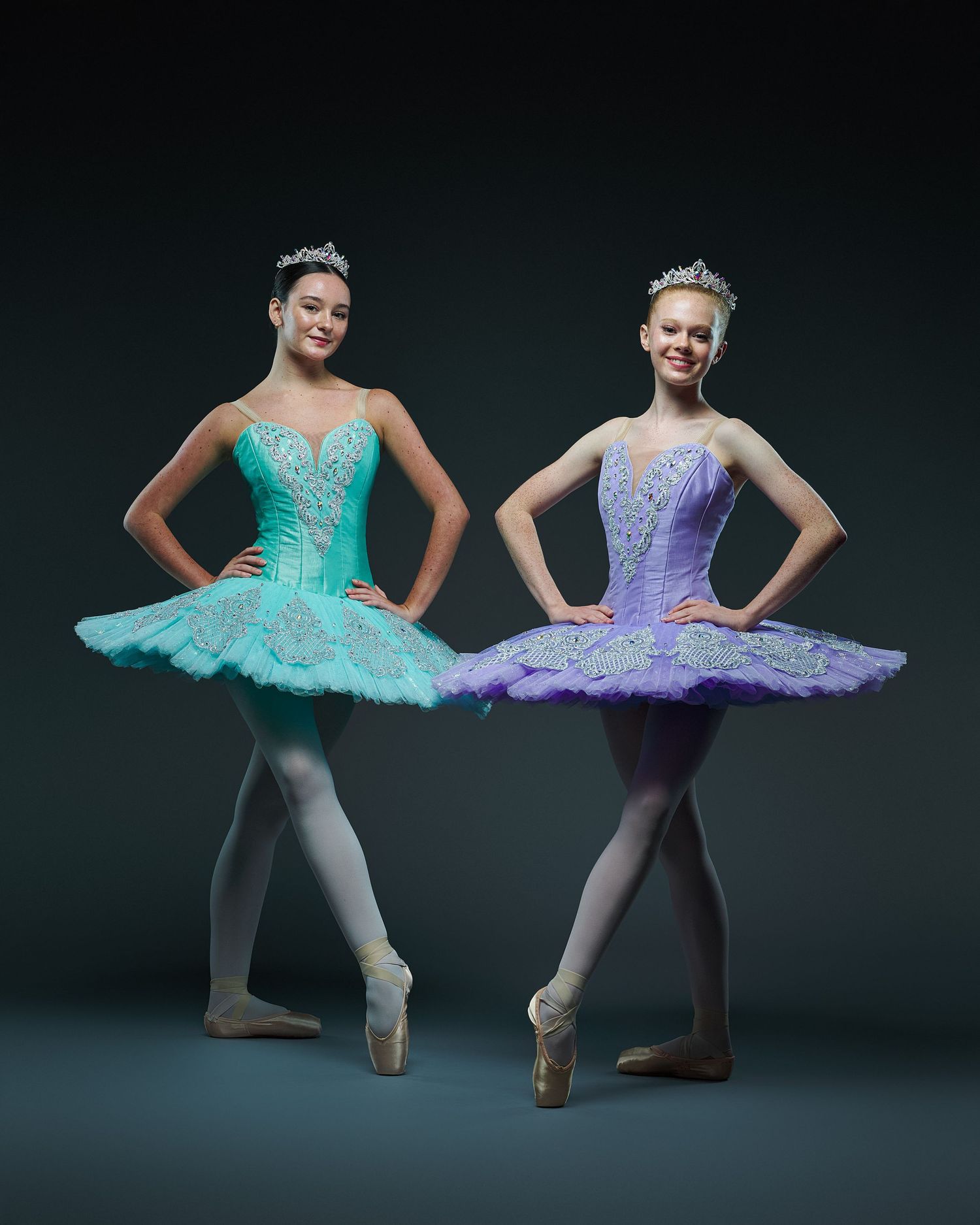 Two dancers pose in mint green and lavender classical tutus against dark background.