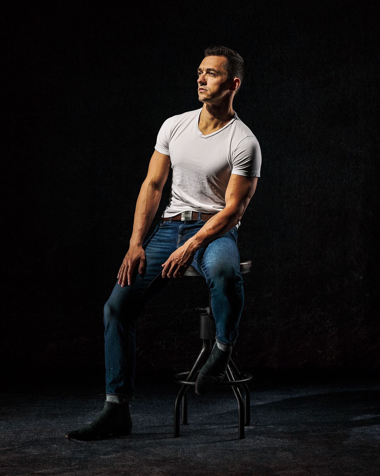 Moody portrait series of person in white t-shirt and jeans seated on chair against dark backdrop.