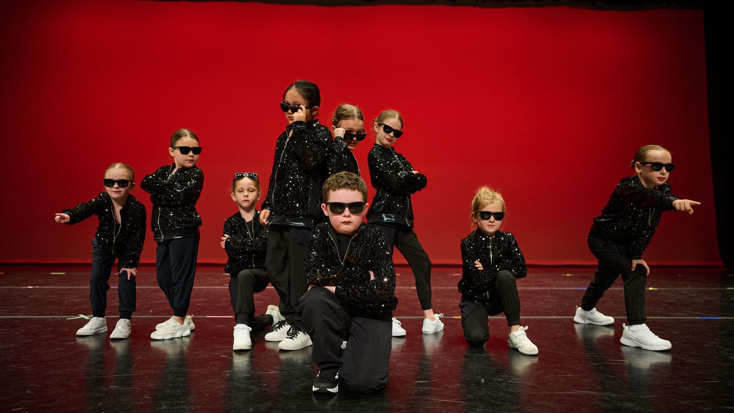 Group of young dancers in black outfits performing coordinated hip-hop routine on red stage.