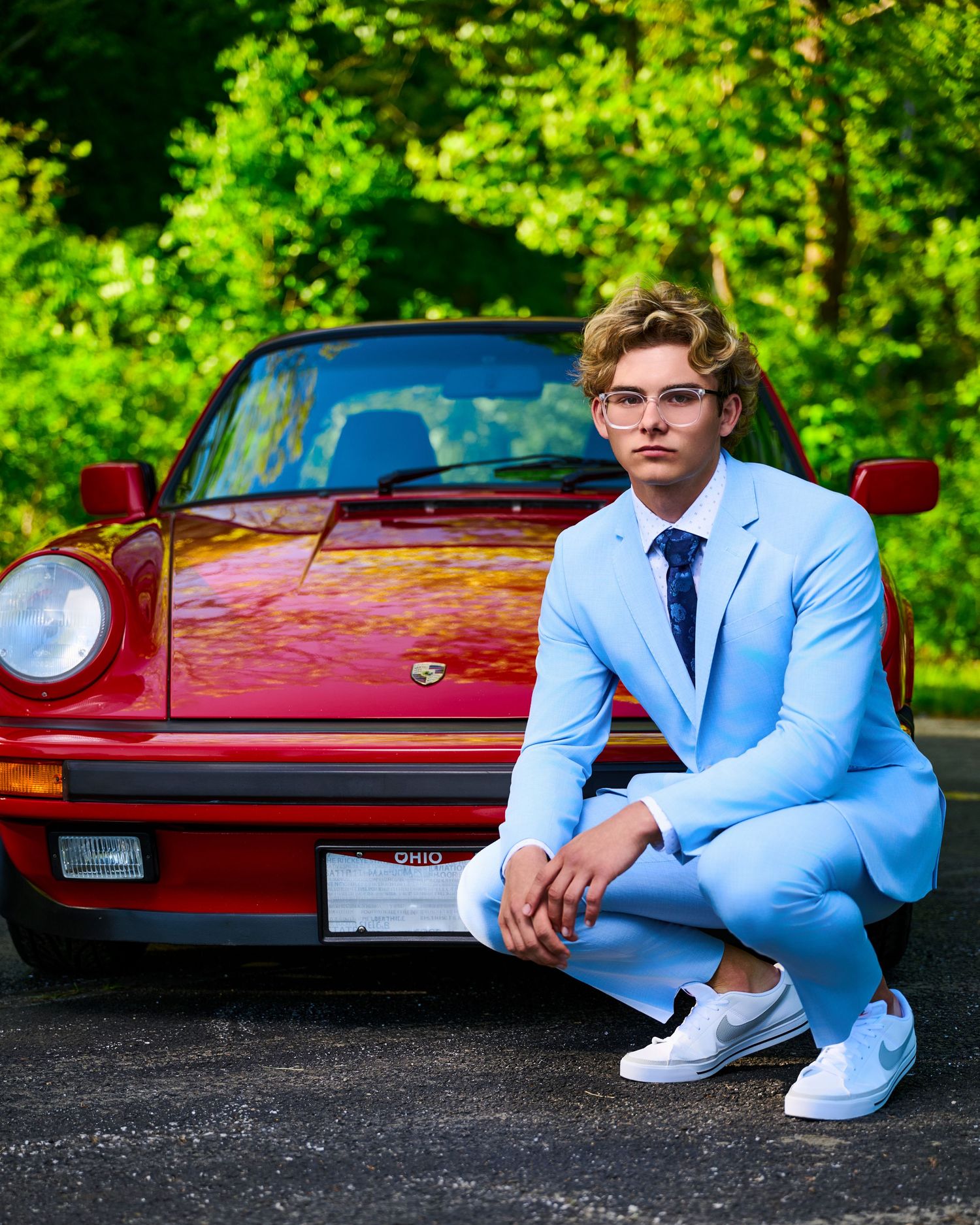 A vintage red Porsche 911 parked in a lush green setting with someone in a light blue suit and white sneakers posing beside it.