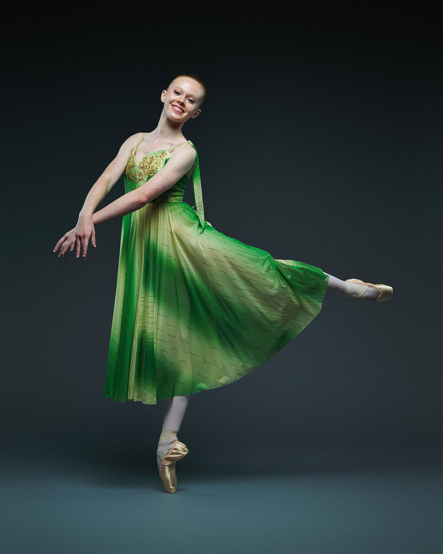 Professional ballet dancer in flowing green dress performs graceful pose on pointe against dark background.