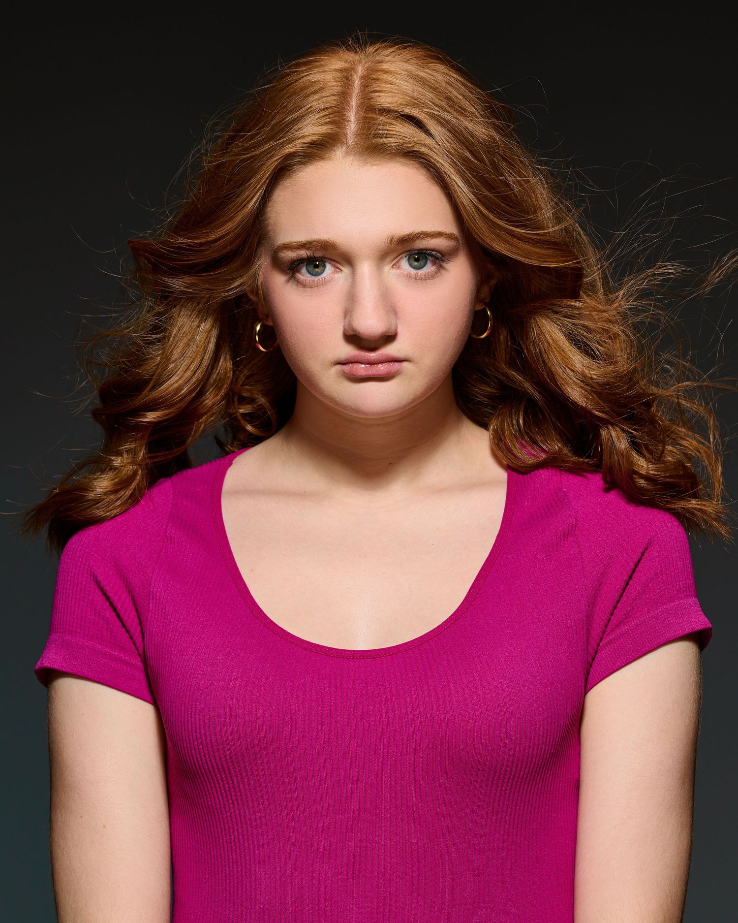 Dramatic portrait with flowing auburn hair wearing magenta top against dark background.