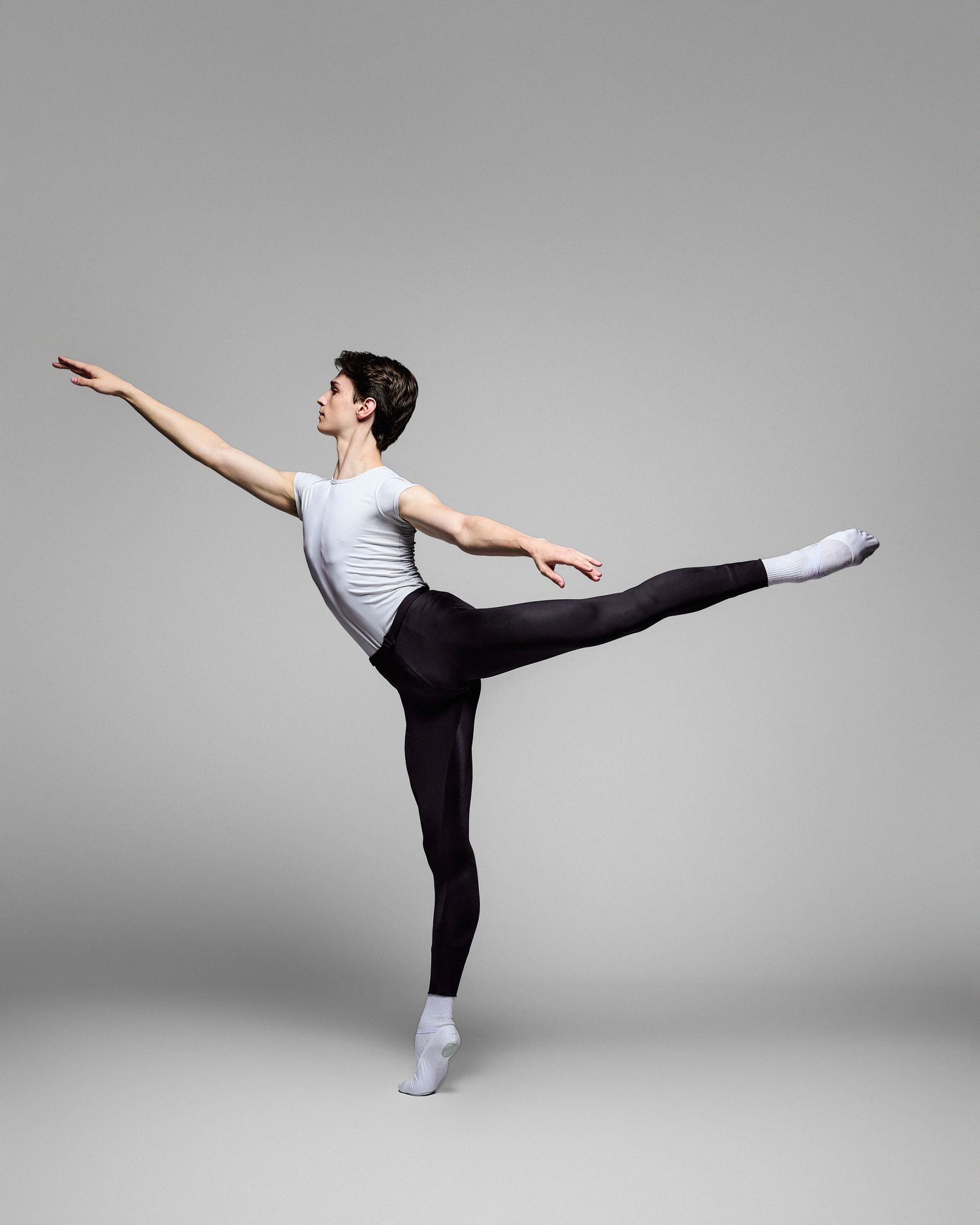 Male ballet dancer in white shirt and black tights performs arabesque against grey backdrop.