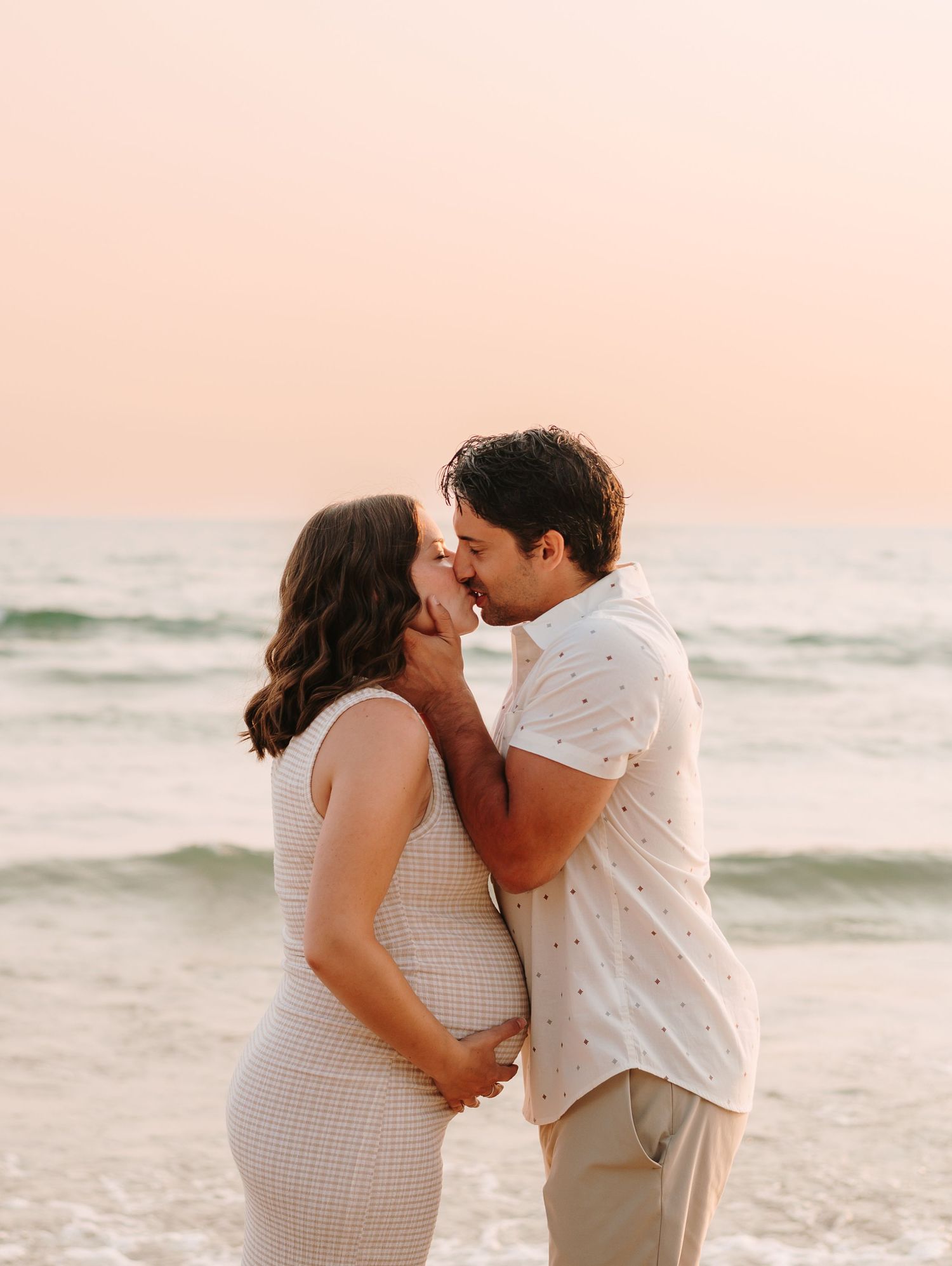 Pregnant couple sharing a romantic moment on a beach at sunset, woman in white fitted dress, man in white shirt.