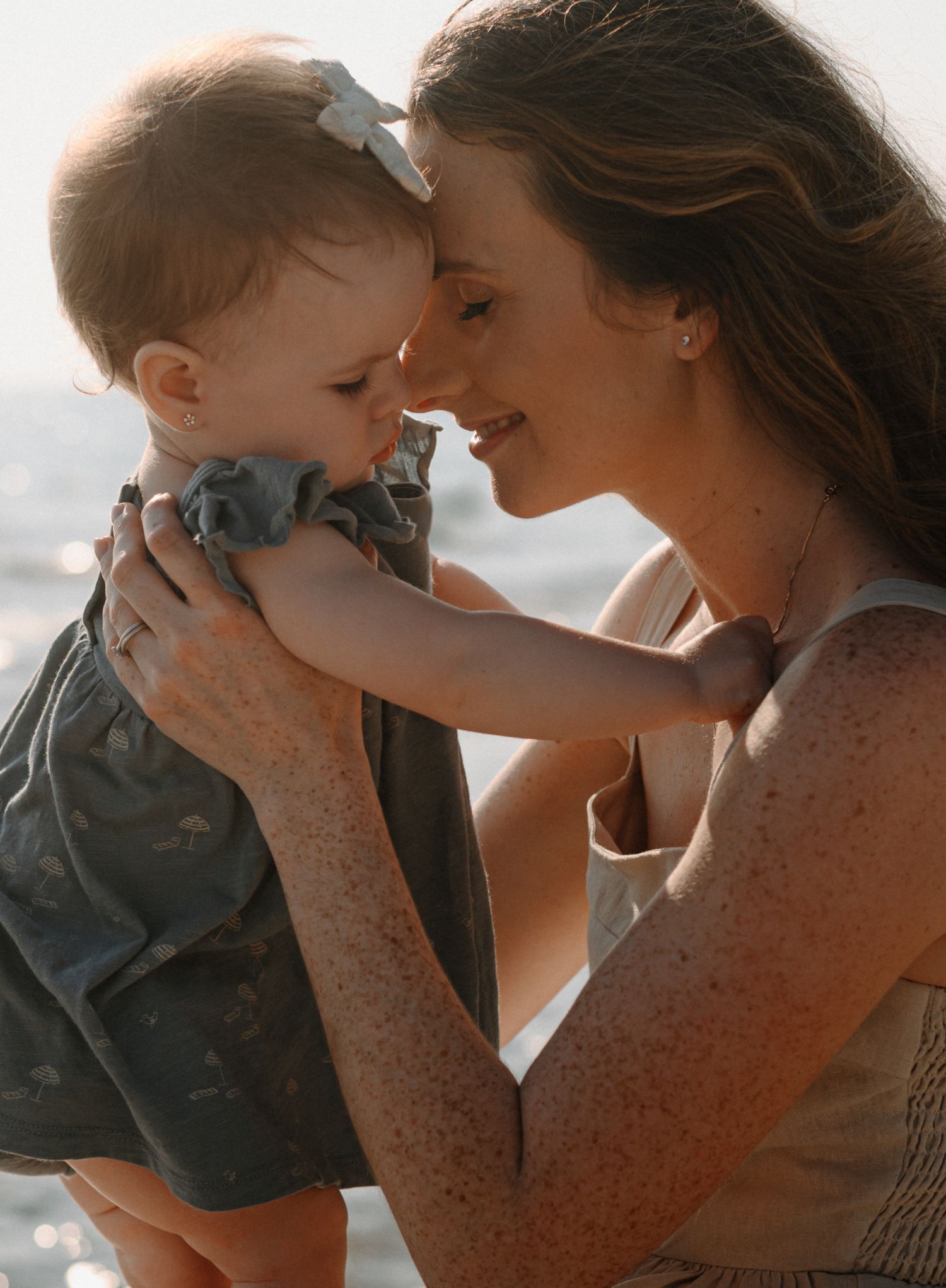 Mother with freckles smiles at baby girl on sunny beach.