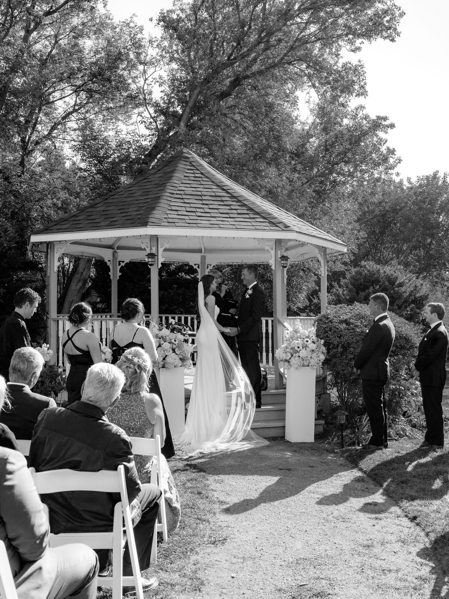 Black and white photo of an outdoor wedding ceremony at a white gazebo surrounded by lush trees.