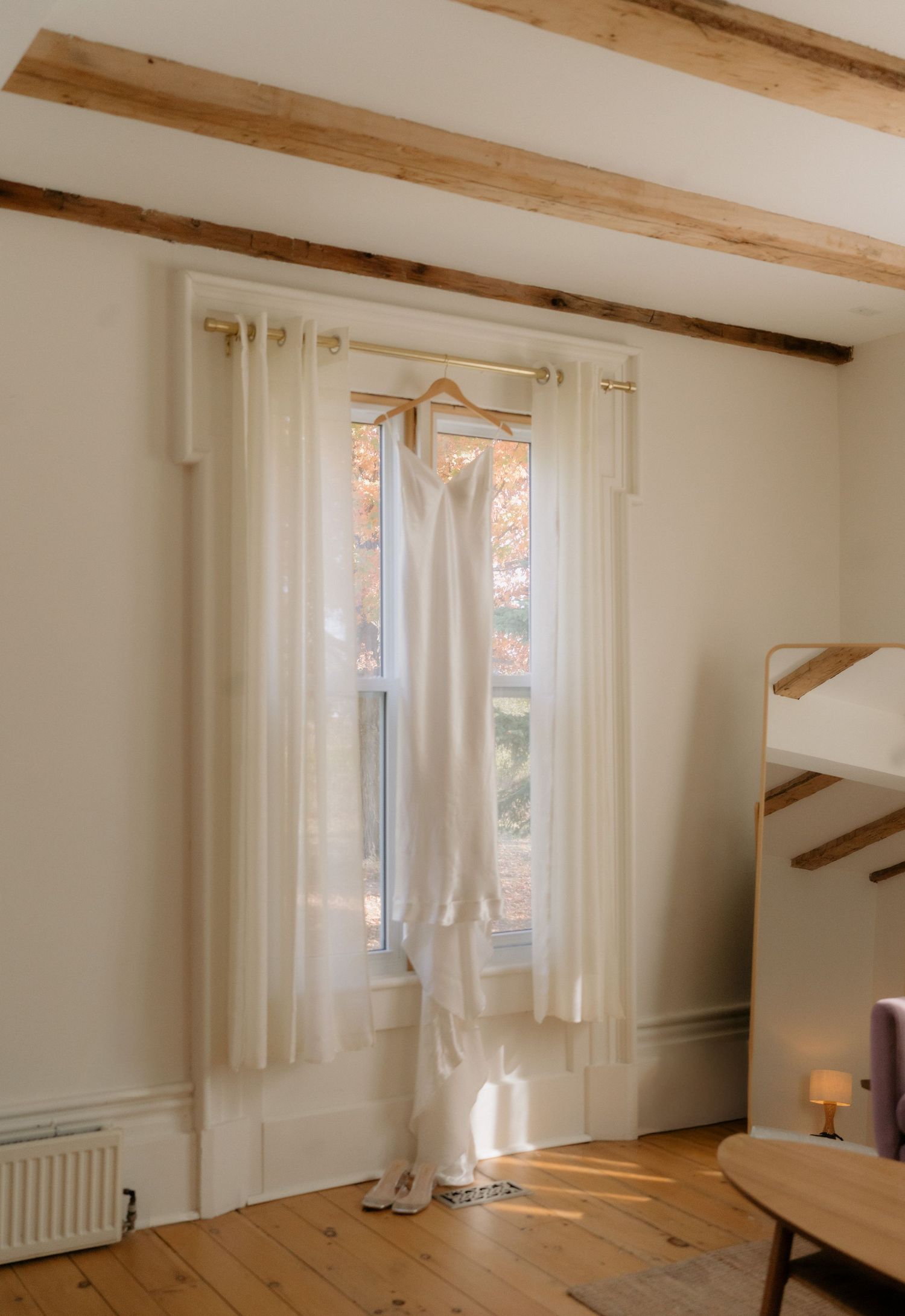 Elegant white wedding dress hanging by a sunlit window in a rustic room with wooden beams.
