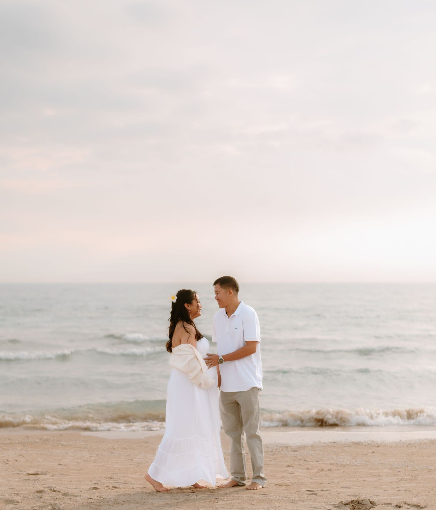 A couple in white outfits shares an intimate moment on a misty beach at sunset.