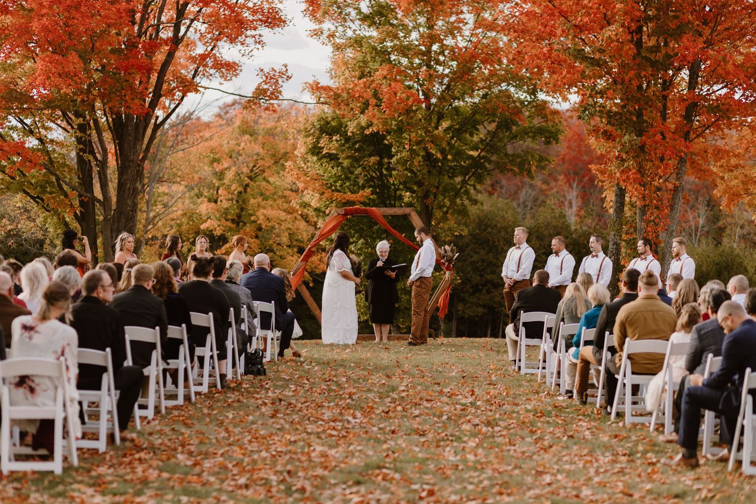 Outdoor autumn wedding ceremony setup with white chairs arranged on fallen leaves under vibrant orange maple trees.
