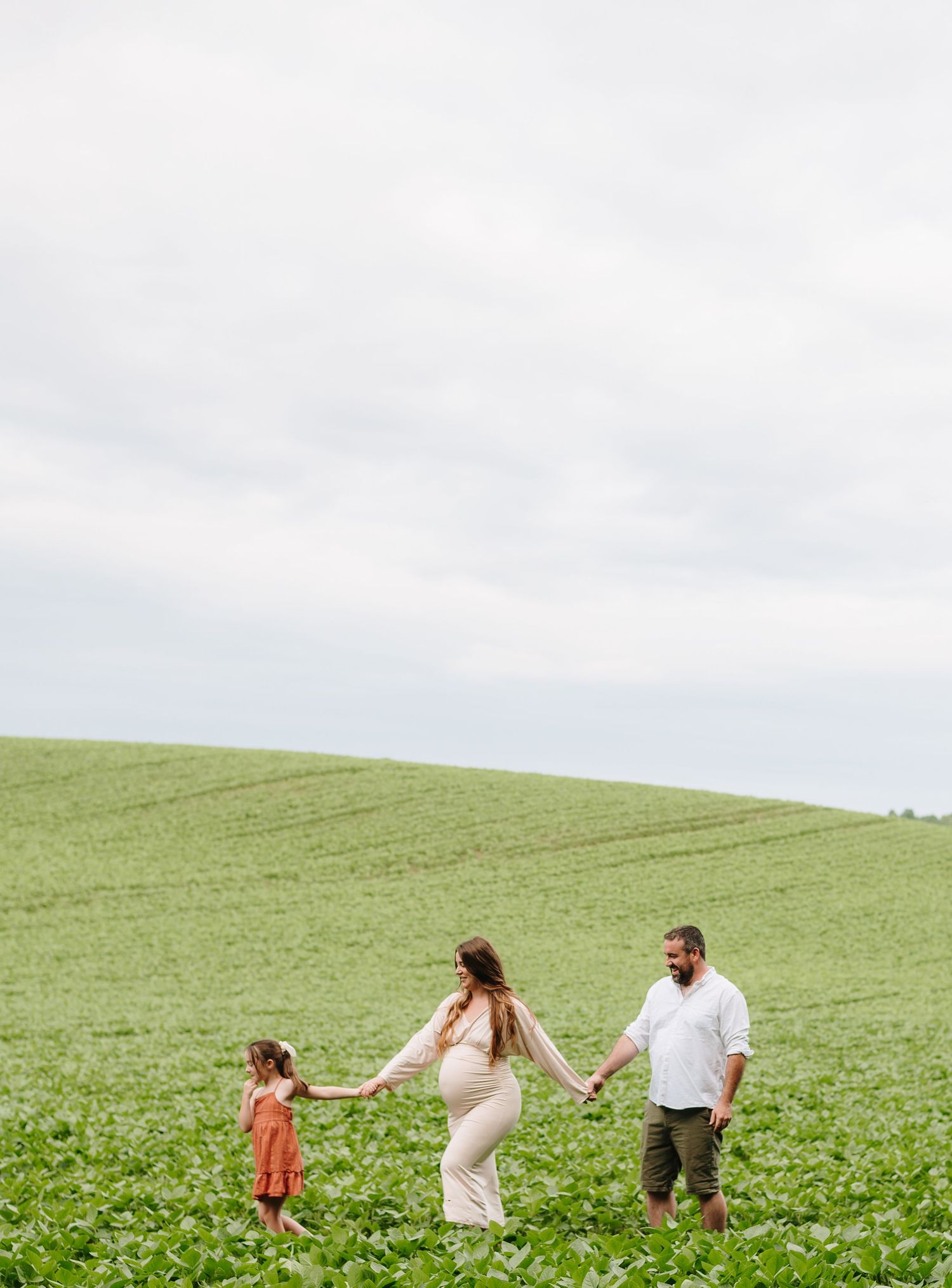 Pregnant woman in beige dress holds hands with husband and daughter in green field.