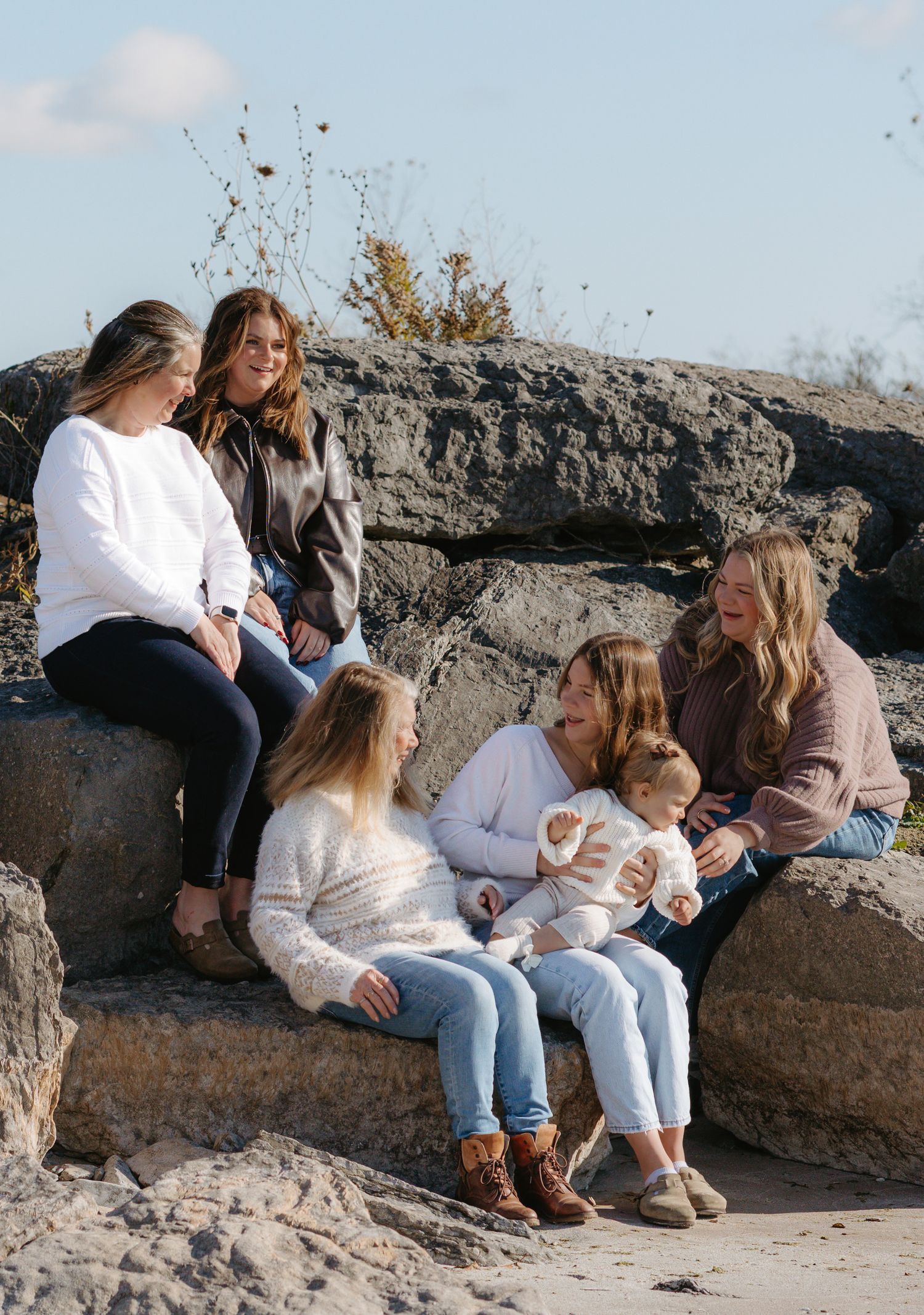 A group sits together on large rocks outdoors during a casual autumn photoshoot in natural lighting  in Prince Edward County