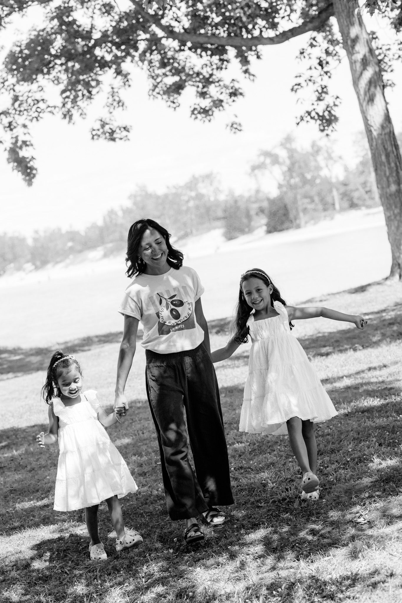 A black and white photo of a parent and two children holding hands while walking under a tree on a sunny day.
