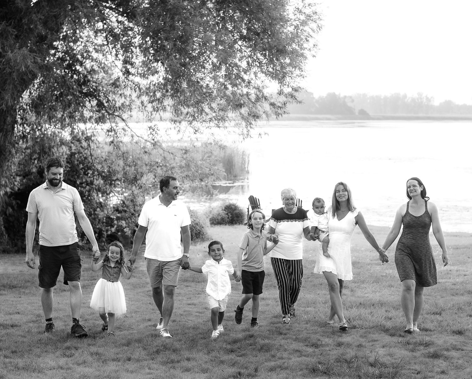 Black and white photo of a large multigenerational family holding hands and walking together near a lake outdoors.