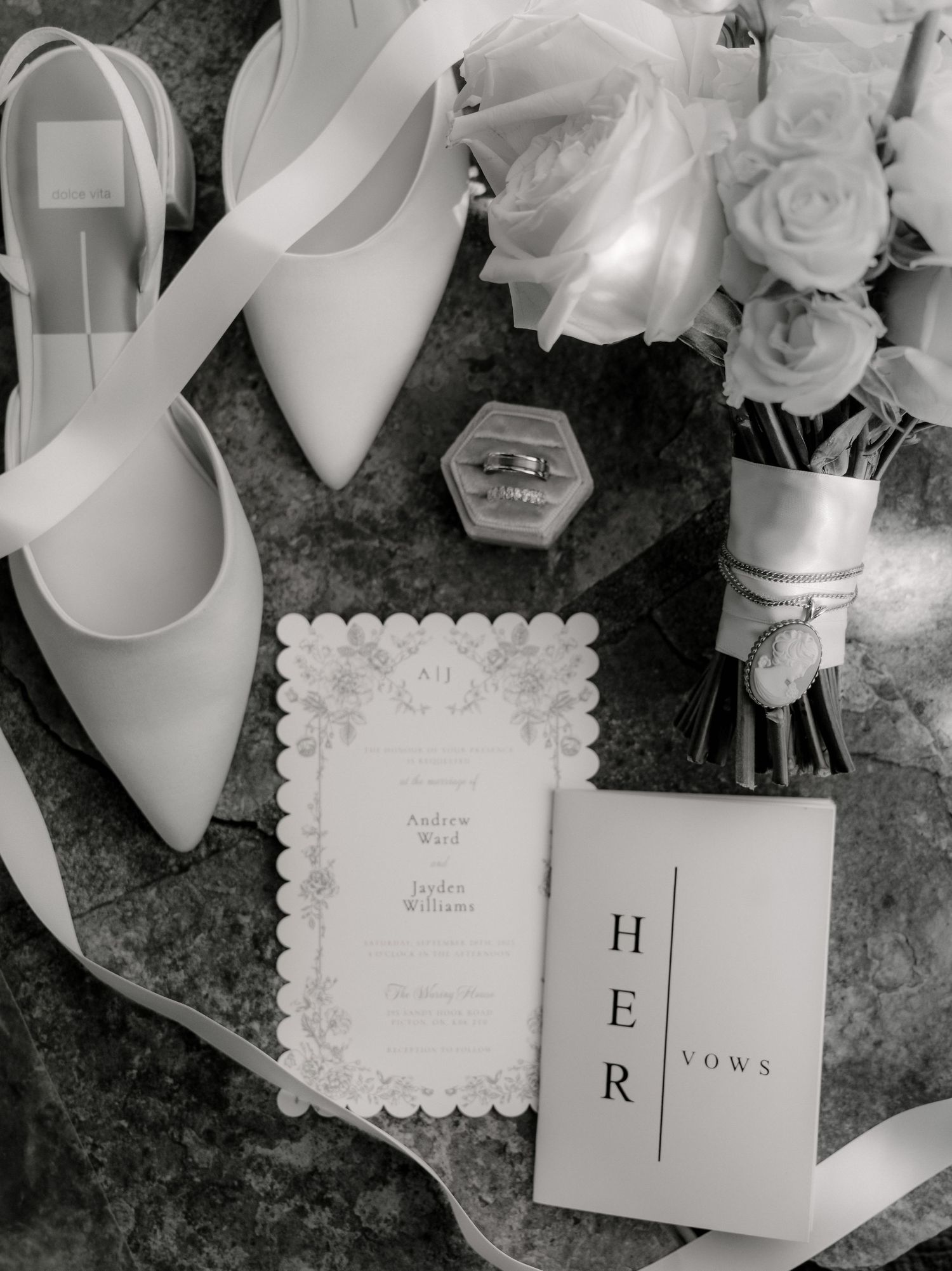 Black and white flat lay of bridal shoes, bouquet, rings, and vow books.