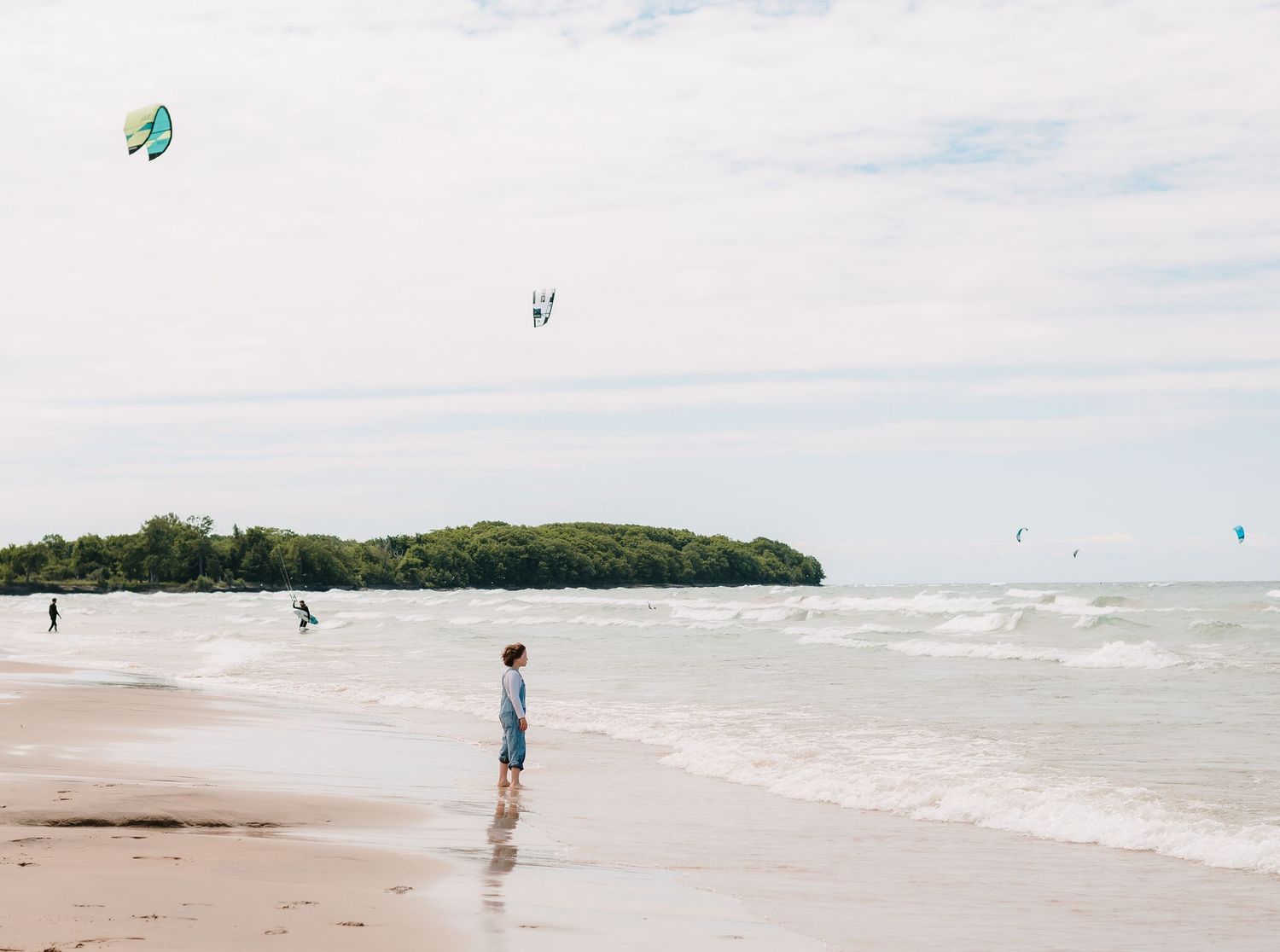 A person stands alone on a sandy beach watching colorful kites soaring above the ocean waves.