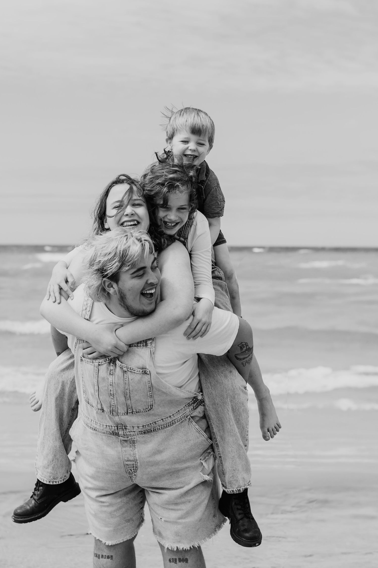 Black and white photo of a man in overalls giving piggyback rides to three laughing children on a beach.