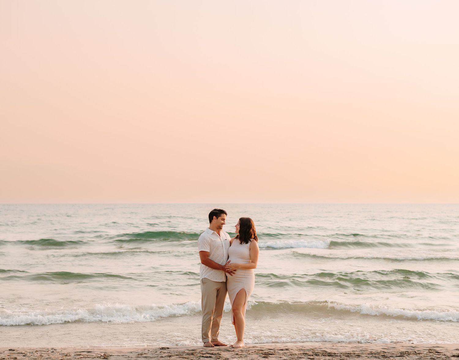 A couple stands together on a beach at sunset with waves rolling in behind them in Prince Edward County
