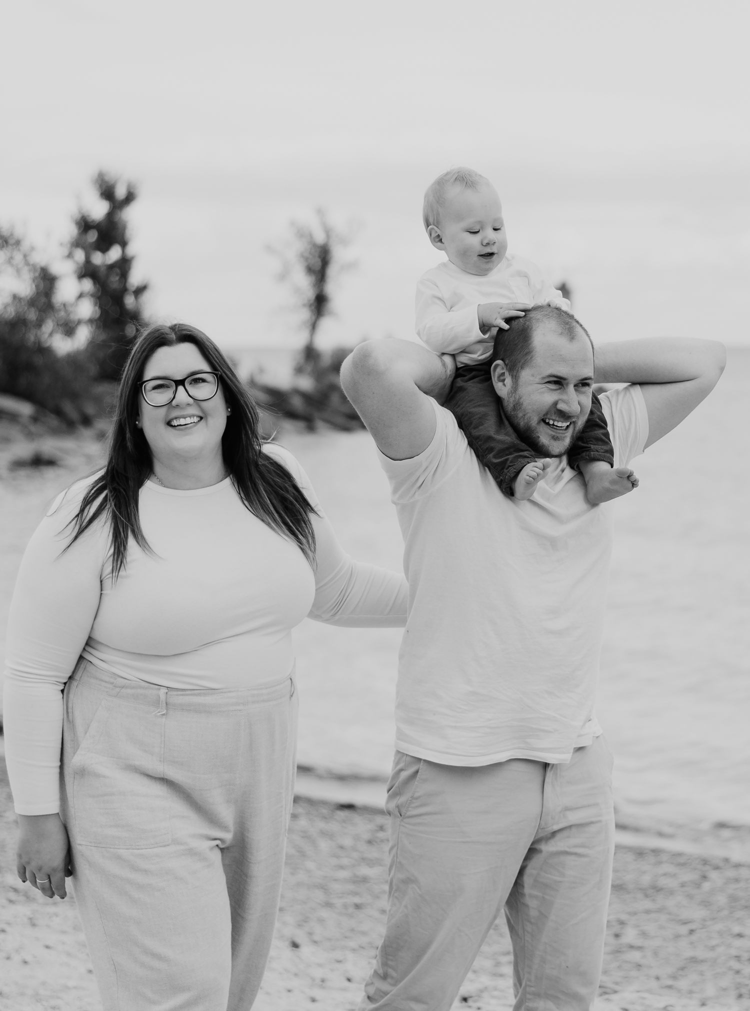 Black and white beach photo of smiling family, mother with glasses, father holding baby on shoulders.
