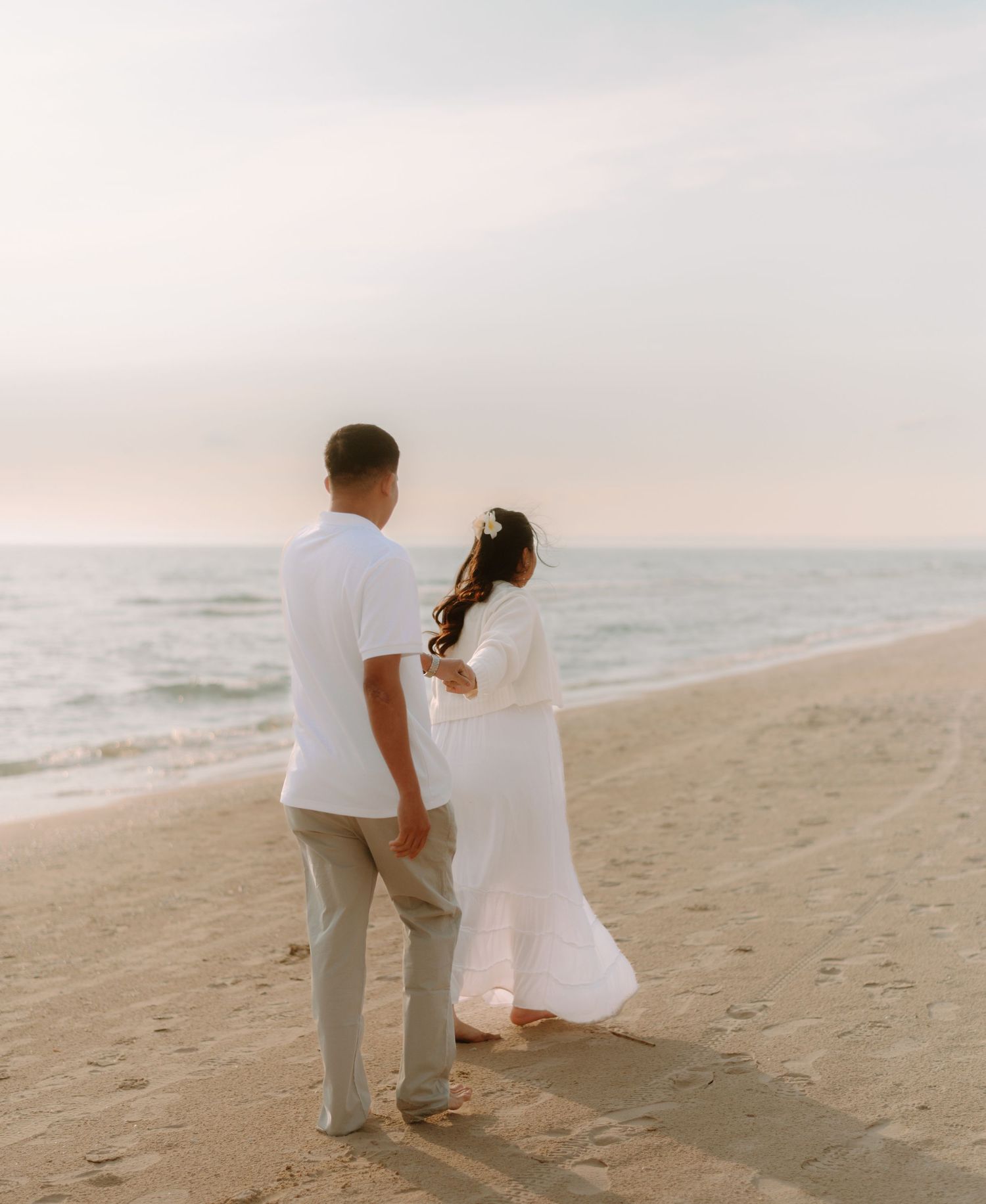 A couple in white outfits walking together along a peaceful beach at sunset.