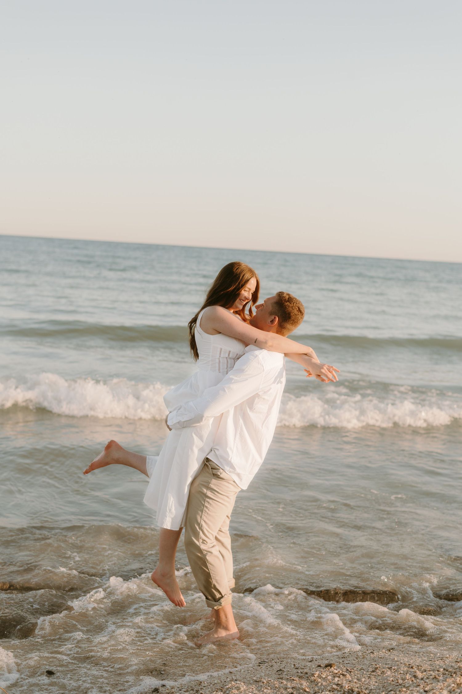 A couple in white clothing dances and embraces playfully at the ocean's edge during sunset.
