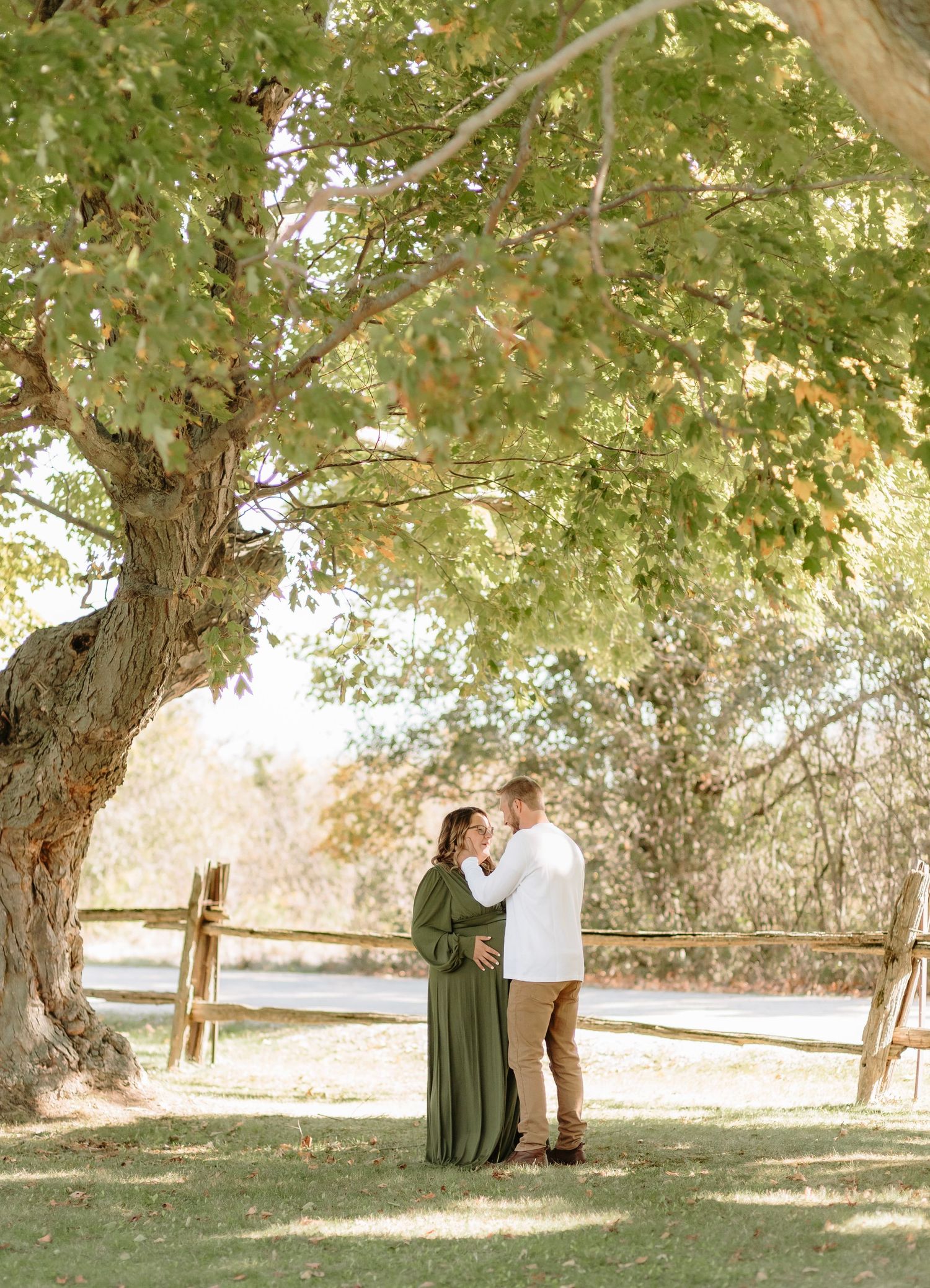 A couple stands intimately under a large tree with sunlight filtering through the leaves.