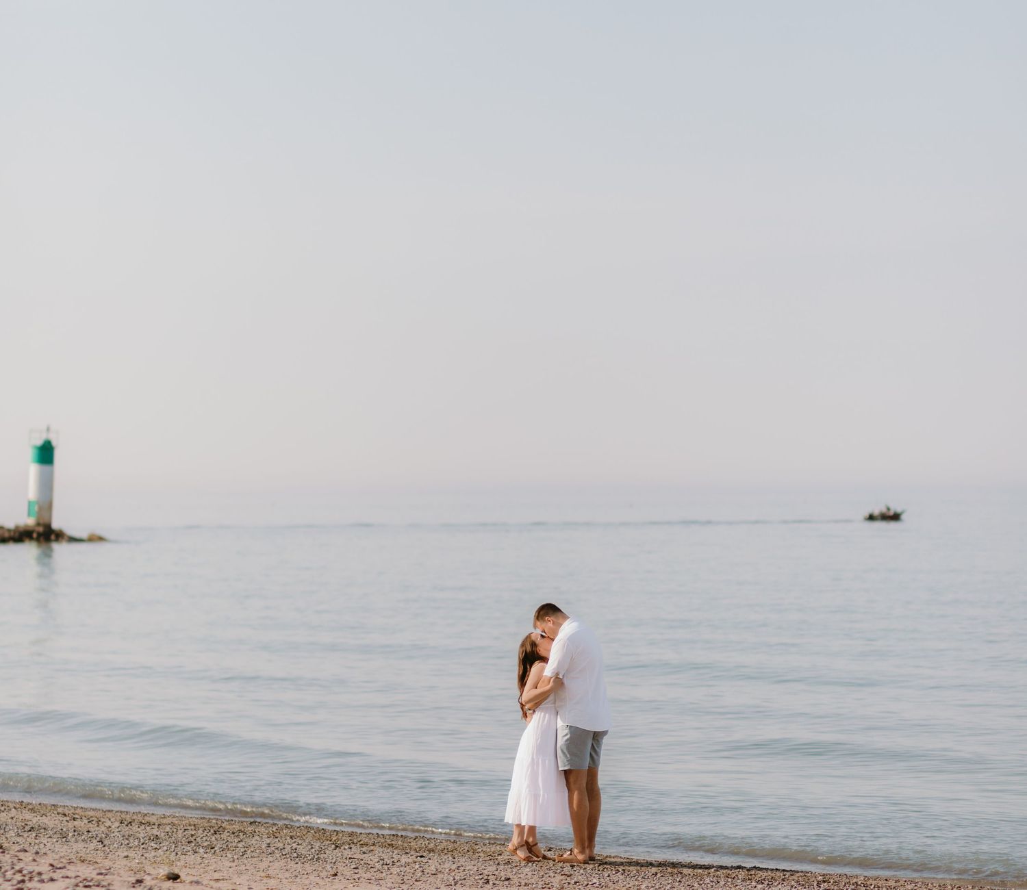 A couple in white attire shares a romantic moment on a misty beach during sunset.