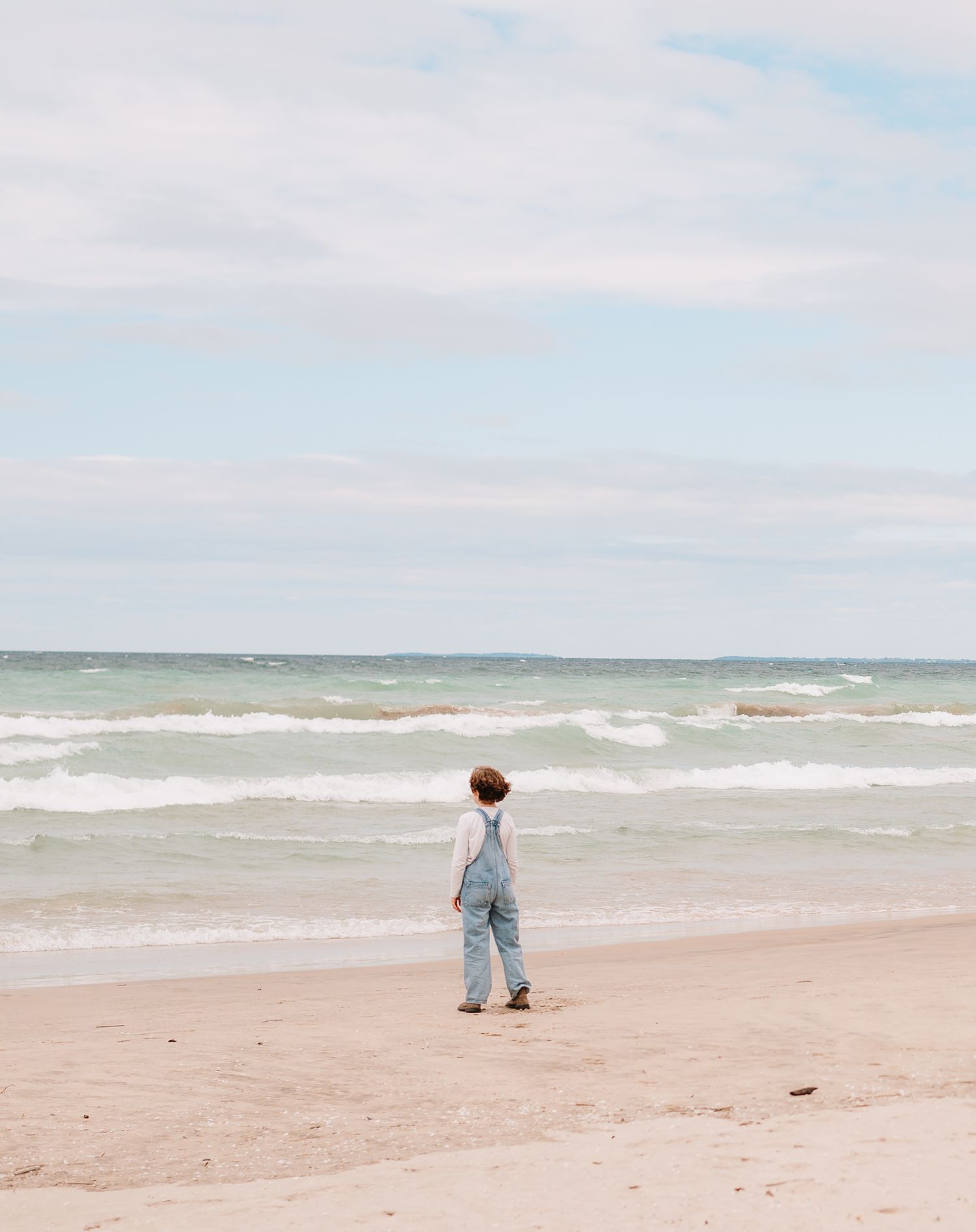 A person in denim overalls stands alone on a peaceful beach looking out at the ocean waves under cloudy skies in Prince Edward County