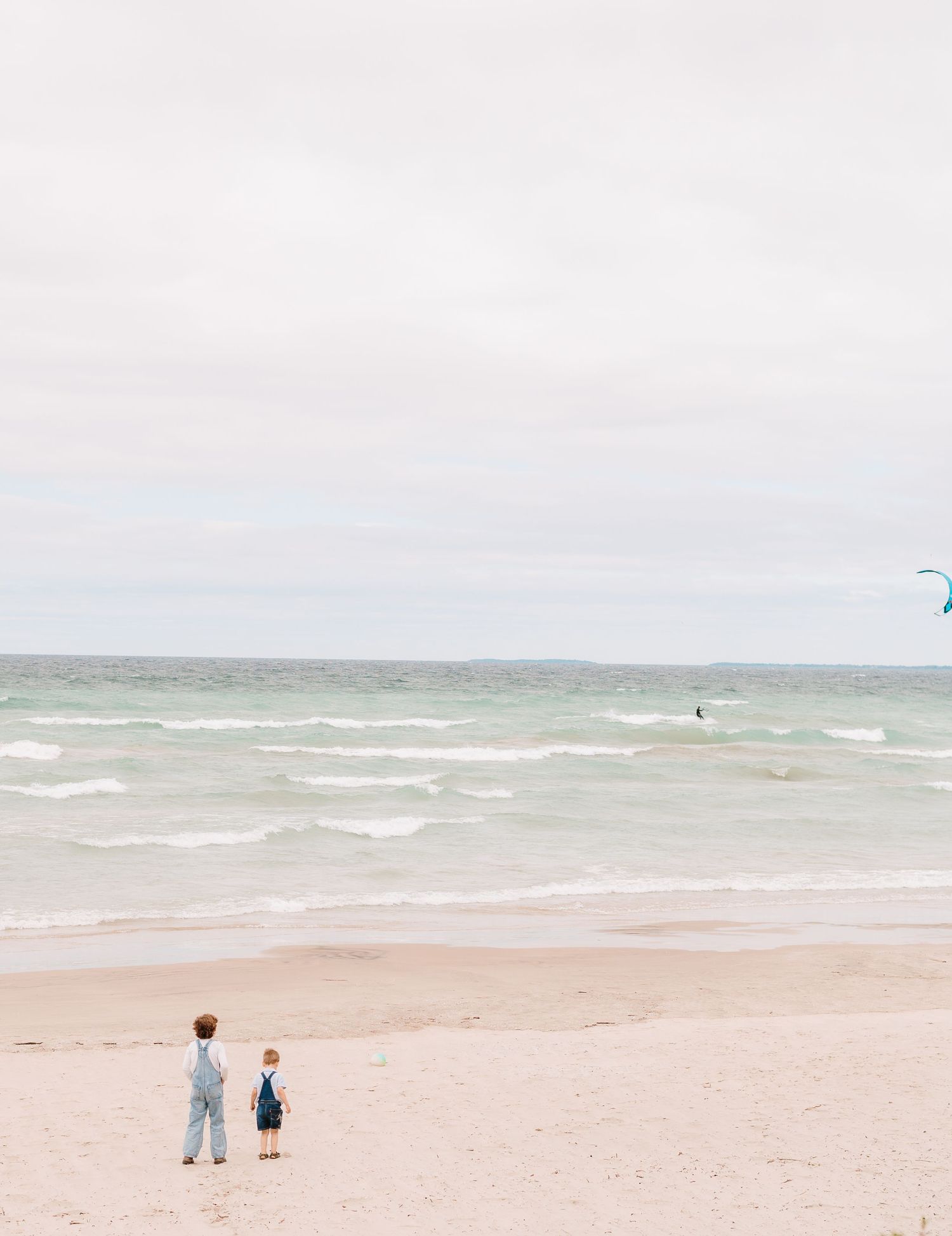 Two young children standing on a sandy beach watching a kite surfer on a cloudy day.