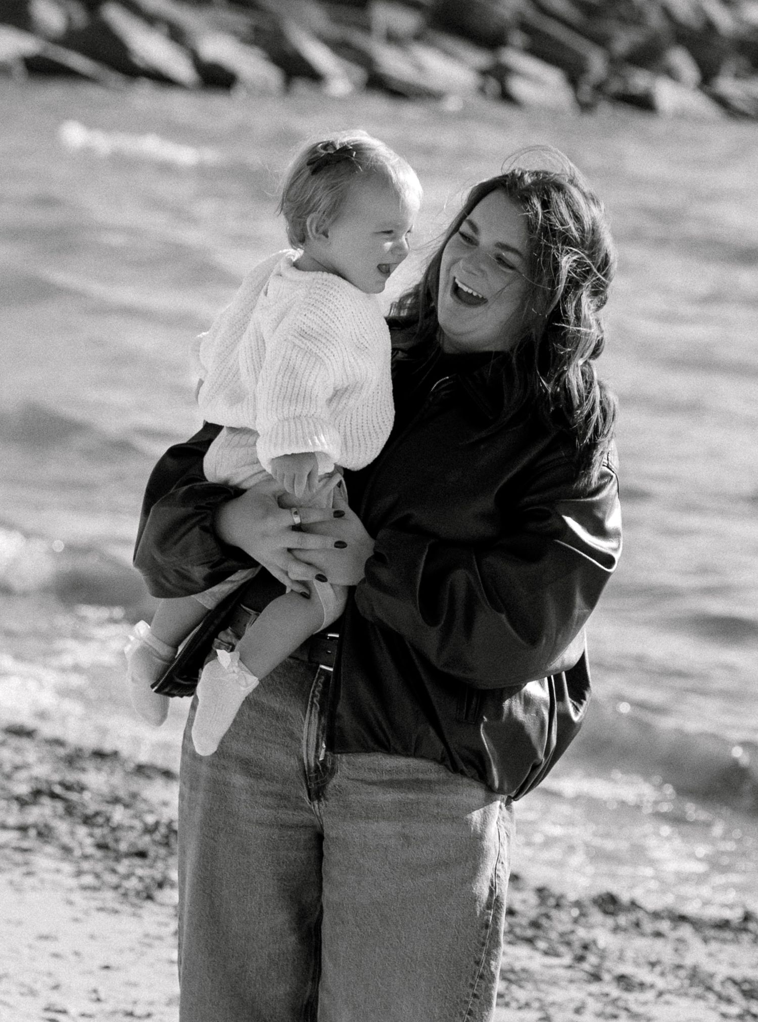 Black and white photo of a laughing mother holding her baby on a rocky beach shoreline.