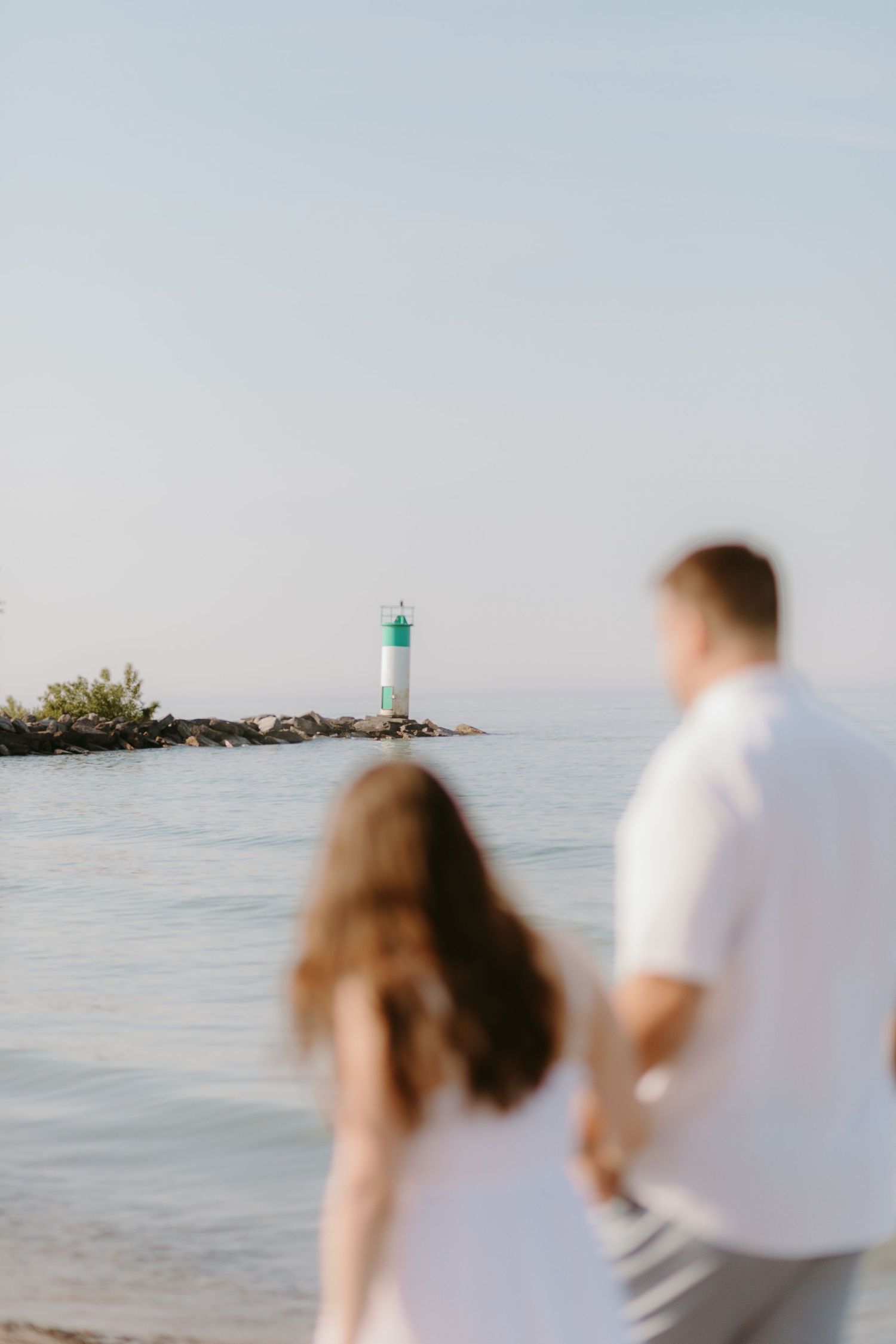 A couple gazes at a green and white lighthouse on the shore during a serene beach day.
