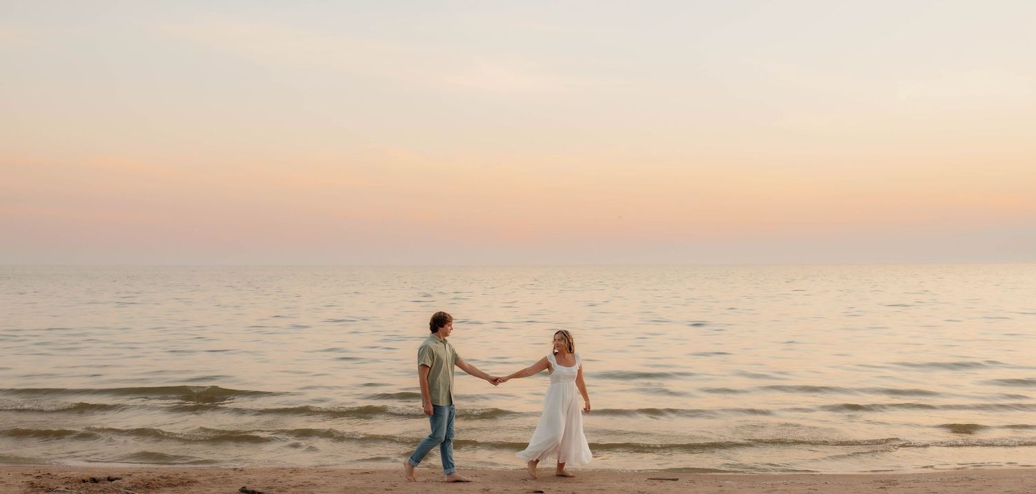 Couple holding hands walking on a sandy beach at sunset with calm ocean waves in the background.