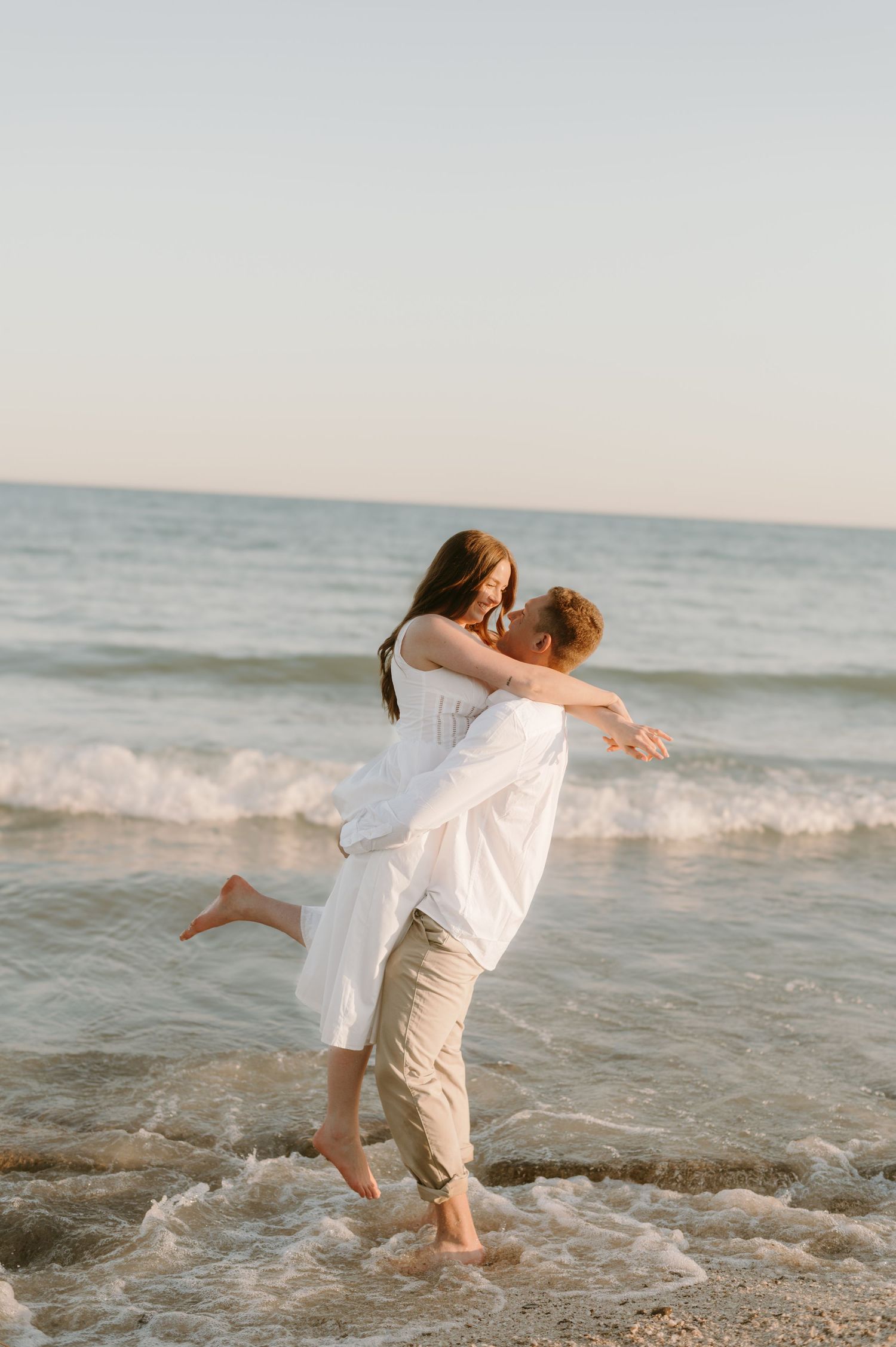 Couple sharing a romantic moment as man lifts woman in white dress at ocean shoreline during golden hour.
