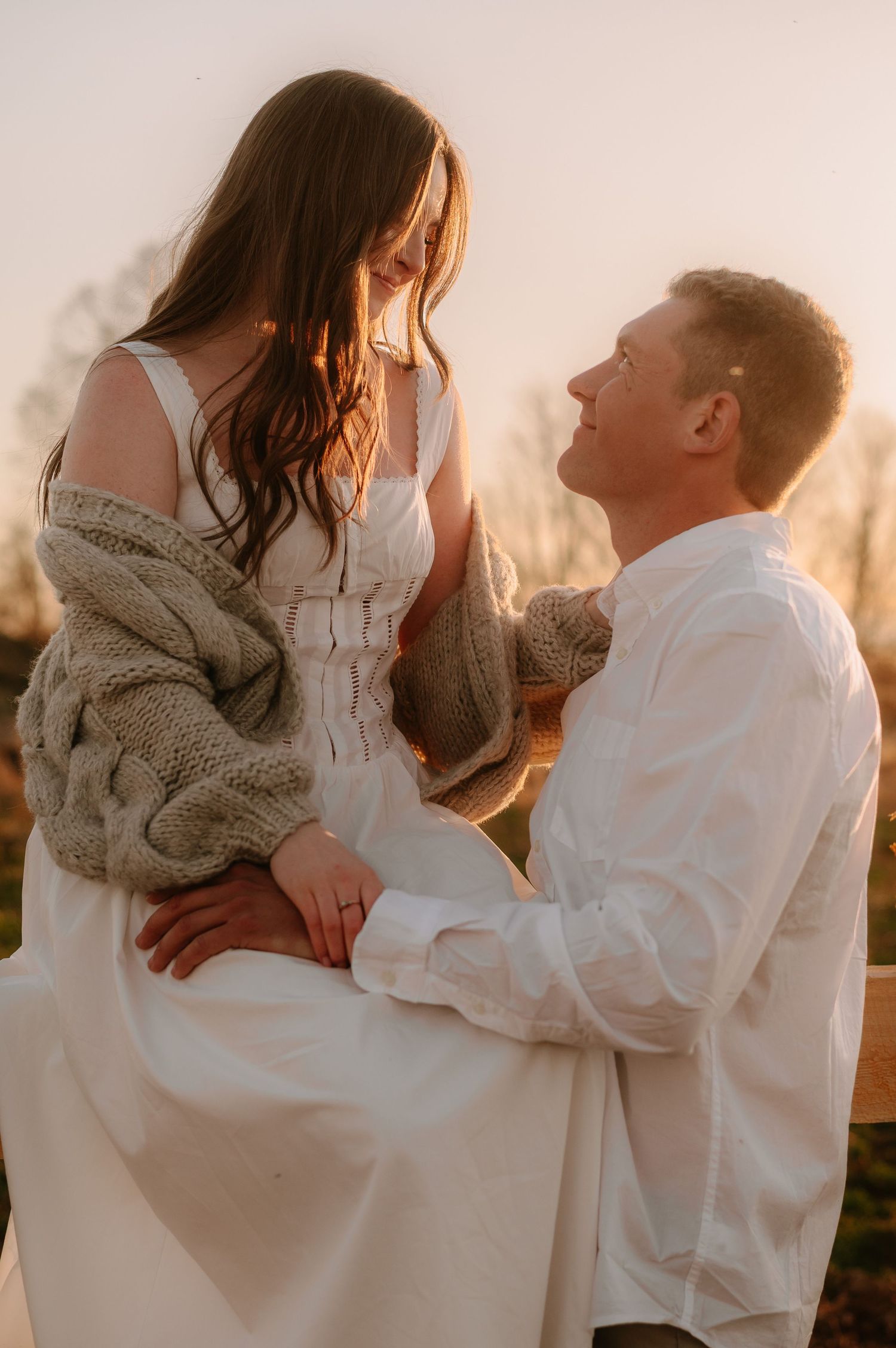 Couple shares a romantic golden hour moment together outdoors, with the woman seated on a fence.