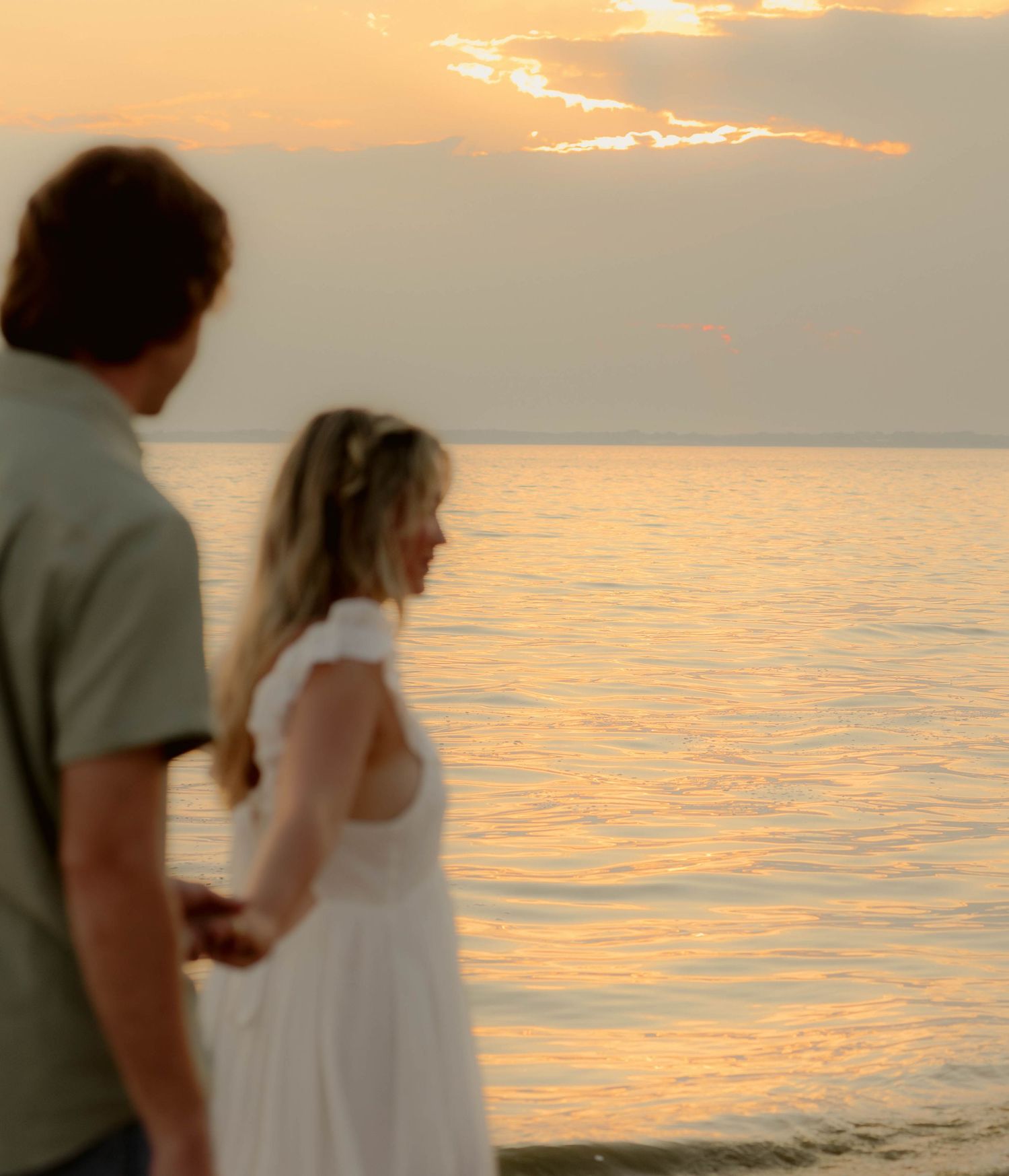 Couple holding hands on a beach at sunset, with golden light reflecting off calm ocean waters.
