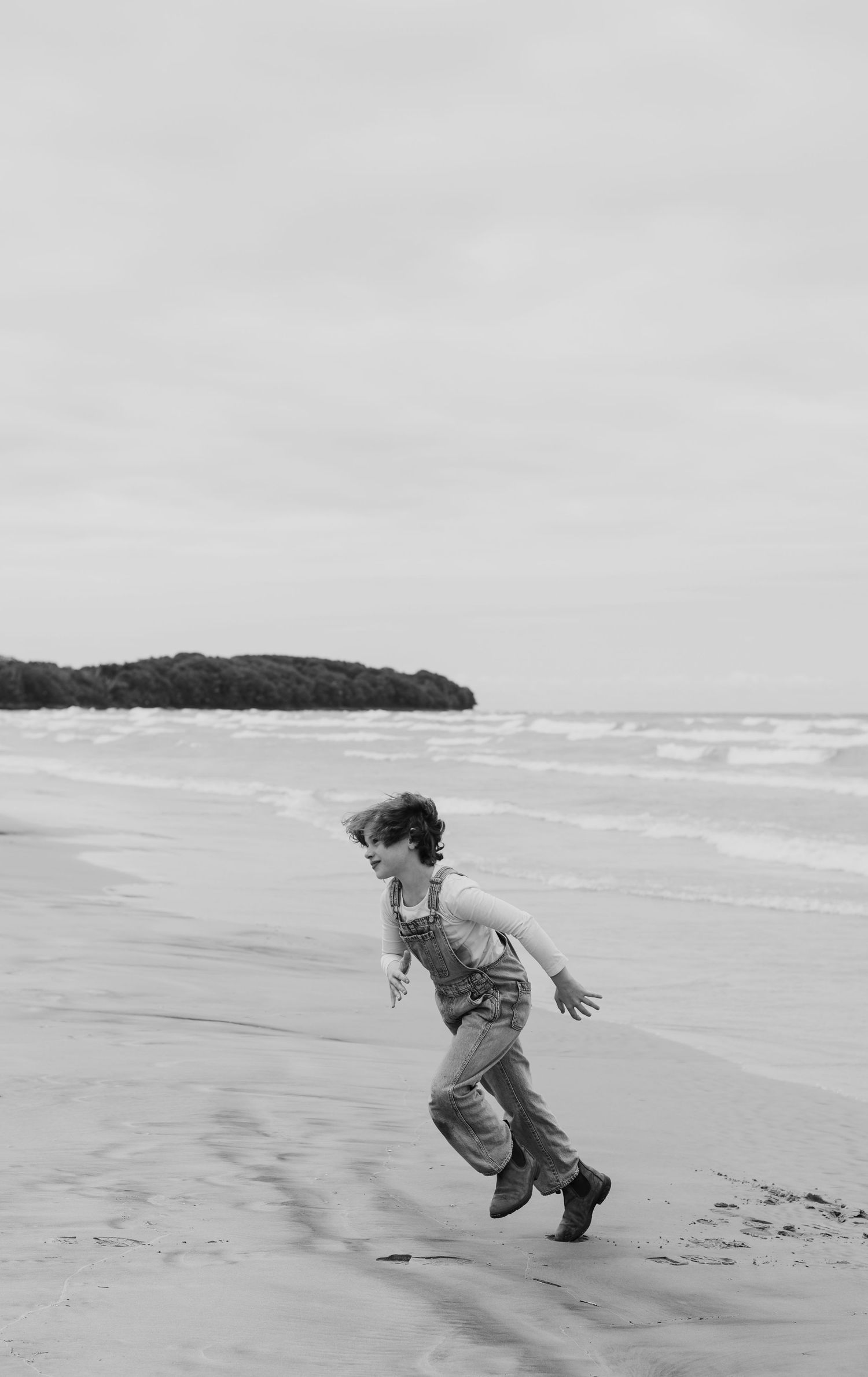 A joyful figure runs along a sandy beach in black and white, with waves and trees visible in the background in Prince Edward County