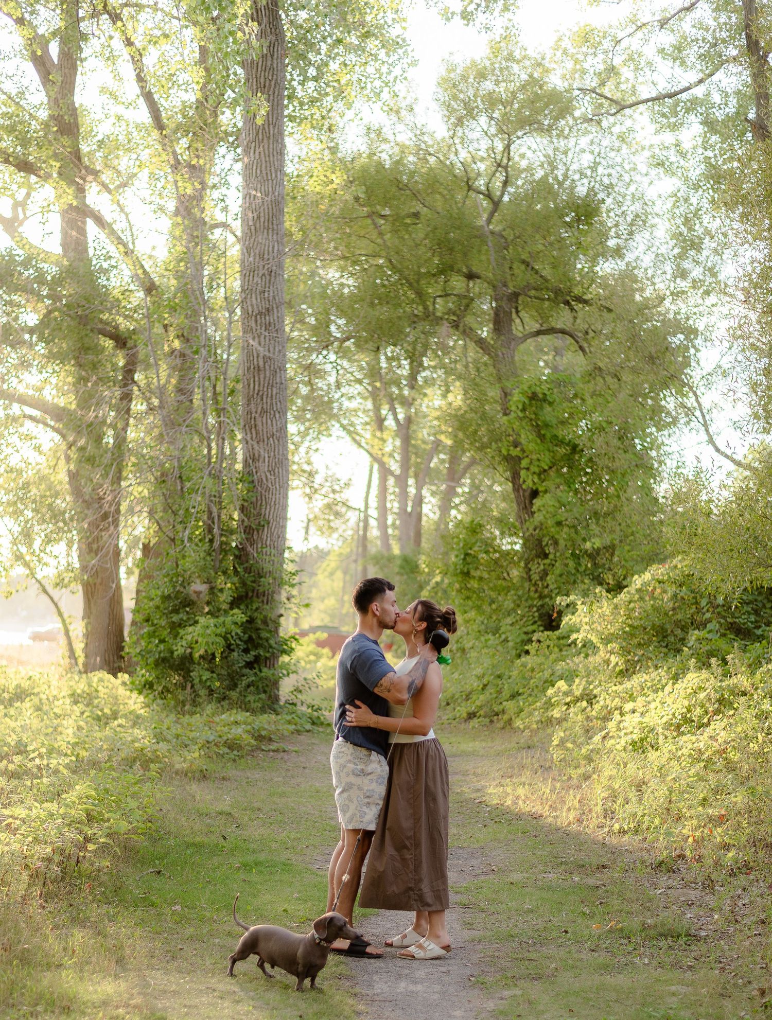 Couple kissing on a sunlit forest path with a dachshund dog at their feet surrounded by tall trees.