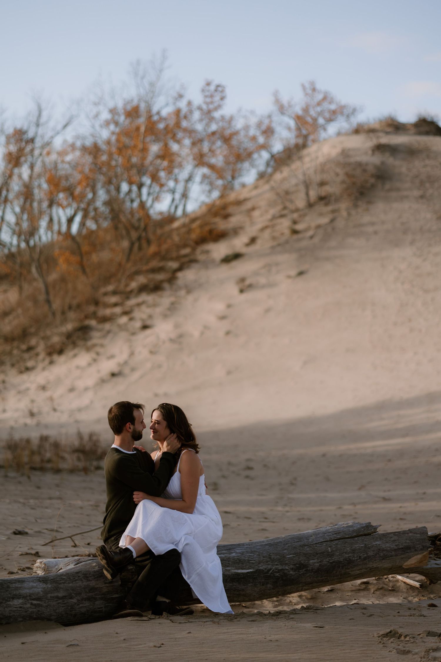 Couple sitting on driftwood log on sandy beach dunes, woman in white dress, man in dark outfit, autumn trees behind.